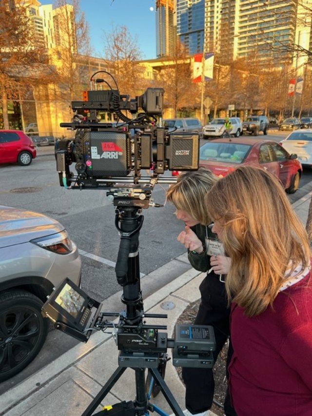 Two women working with a professional video camera mounted on a tripod on a city street with parked cars and tall buildings in the background.