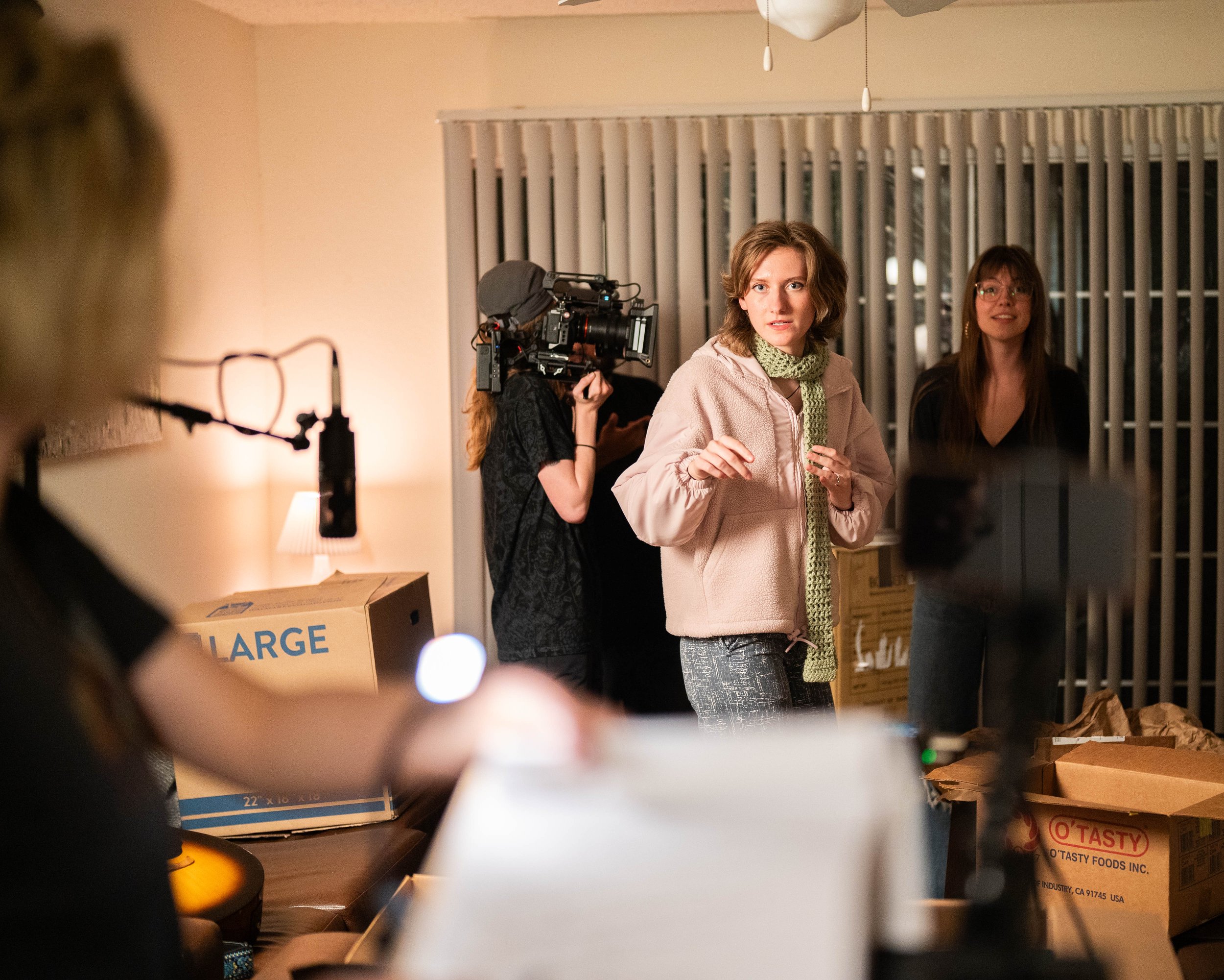 A woman with short brown hair in a pink hoodie with a green scarf appears to speak or present, with two women and a crew member with a camera behind her. The scene takes place in a room with moving boxes.