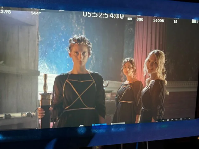 Three women dressed in black standing in a room with elegant decor, backlit by a window with a view outside, and draped curtains.