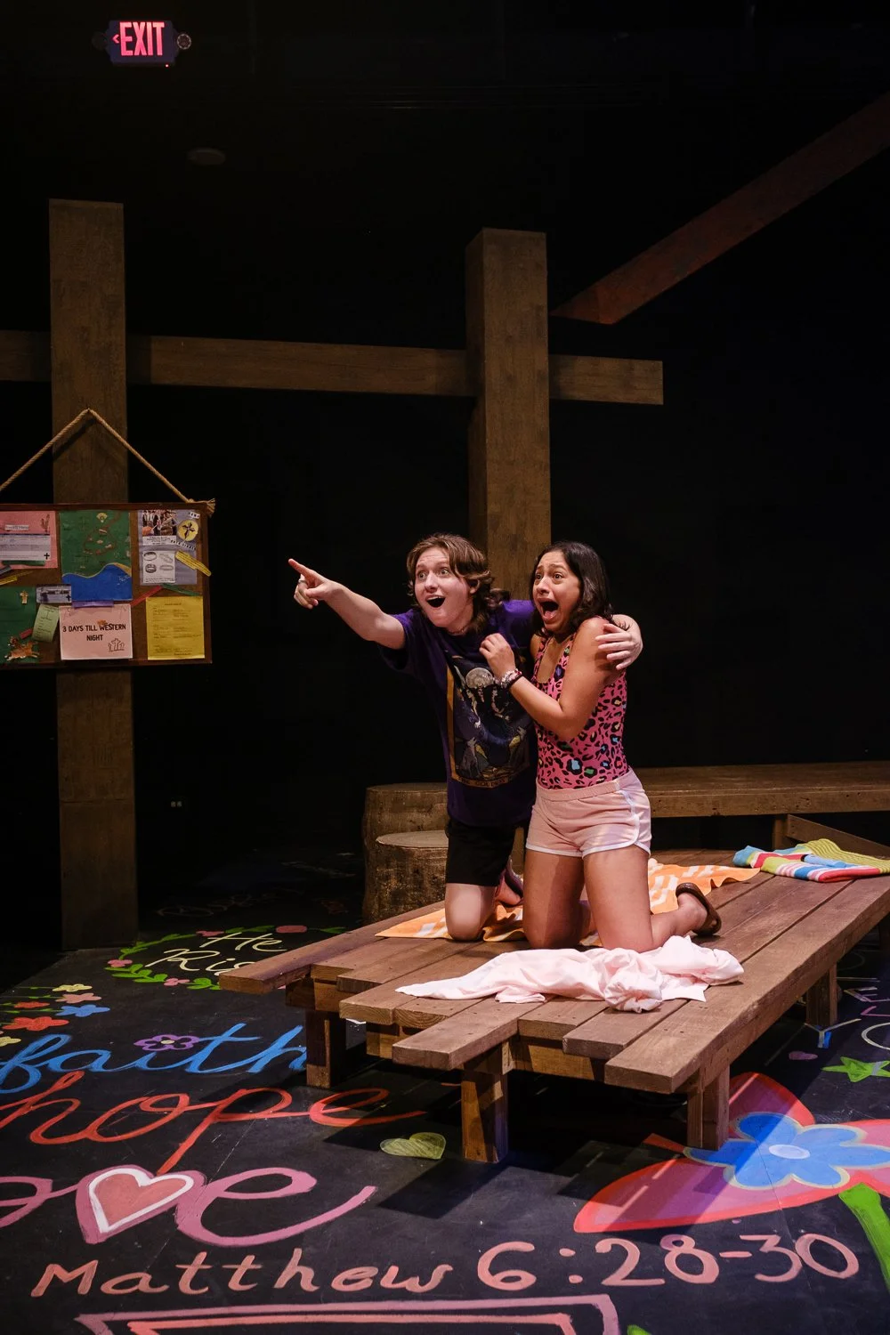 Two women kneeling on a wooden platform, one pointing excitedly while the other looks surprised, in a room with a colorful chalkboard floor and a bulletin board in the background.