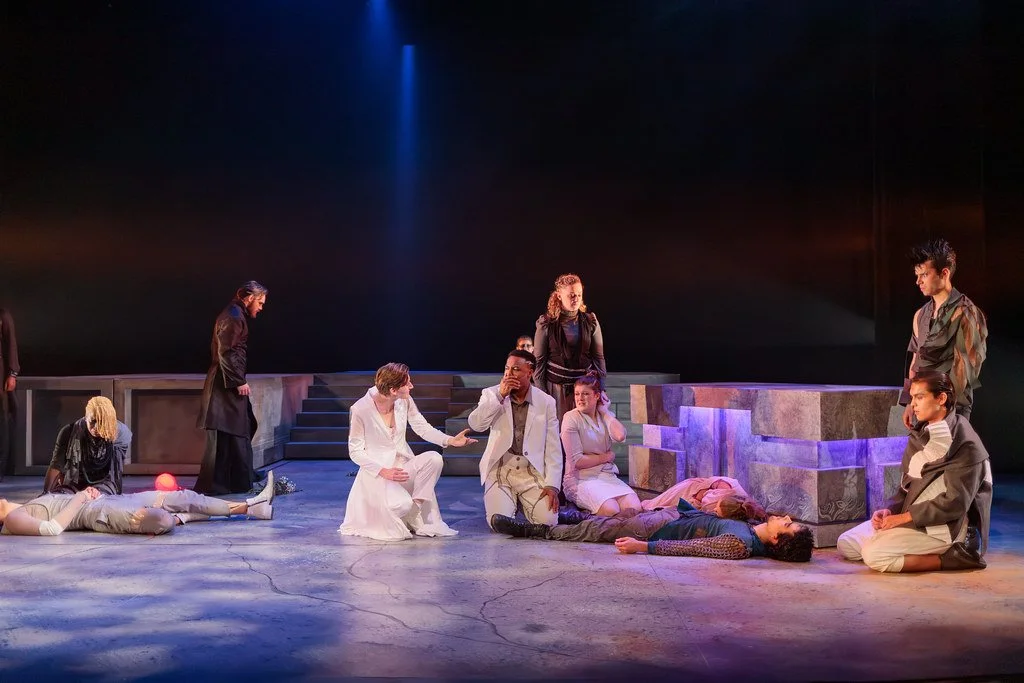 Theatrical performance with actors on stage, some lying on the ground, others sitting or kneeling, illuminated by blue and orange stage lighting.