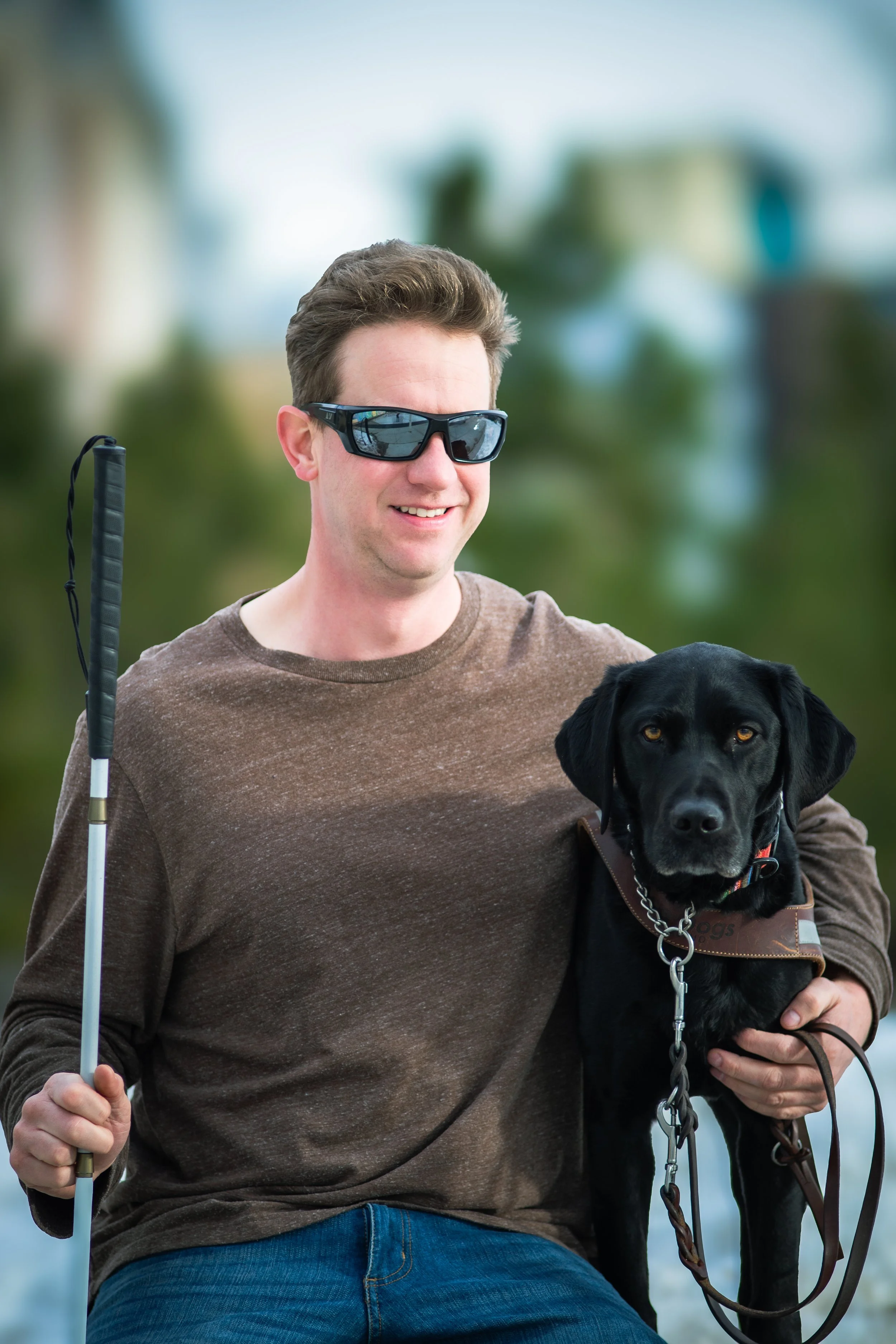 David and his black lab guide dog, Trooper. David wearing sungllasses is holding a white guide cane in his right hand and Trooper in his left. Sitting outside with green trees in the background