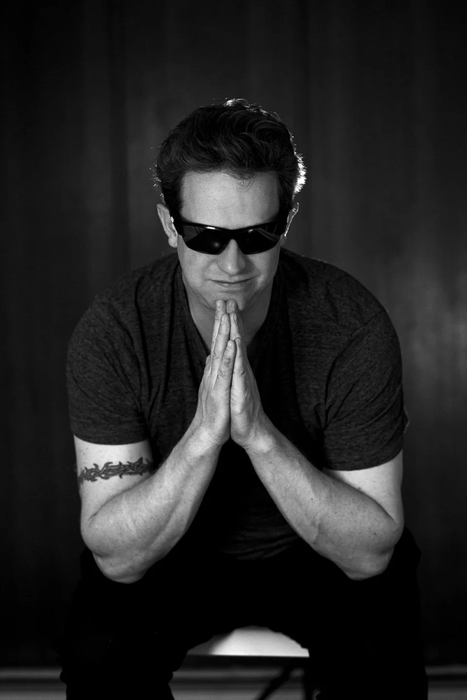 Black and white photo of David wearing sunglasses, with hands pressed together in a prayer-like gesture, head bowed, and a calm expression.