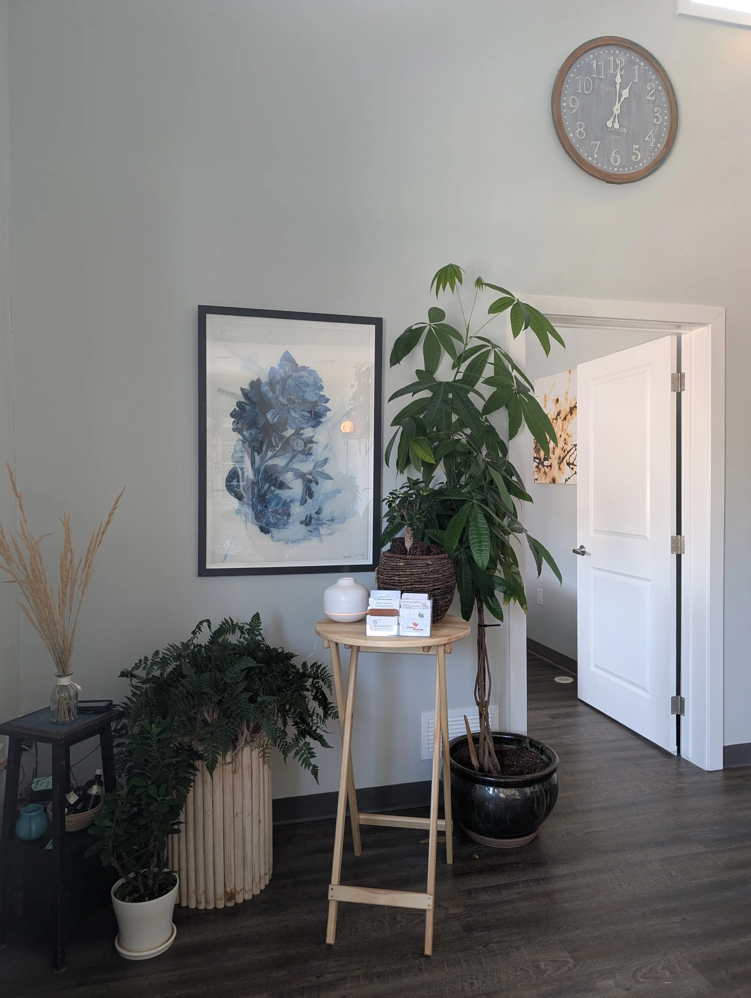 Common area of the Collective with a framed blue watercolour art piece, a tall indoor plant, and smaller potted plants on the floor. A small round table with business cards and a small plant on it.