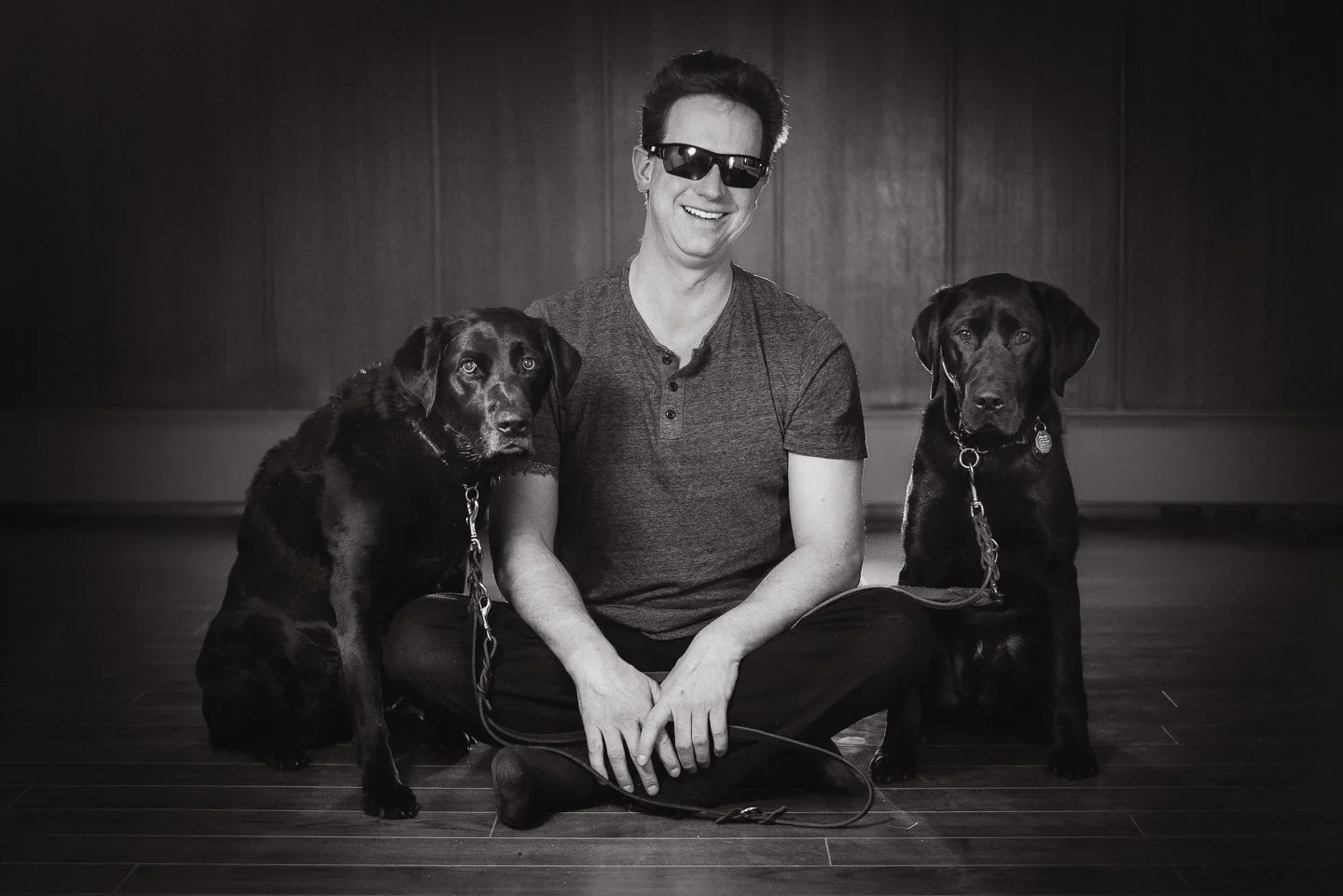 Black & white image of David and his black lab service dogs, McCoy and Trooper sitting on the floor in a room with wooden walls and floor. David smiling with a dog sitting on eithier side,