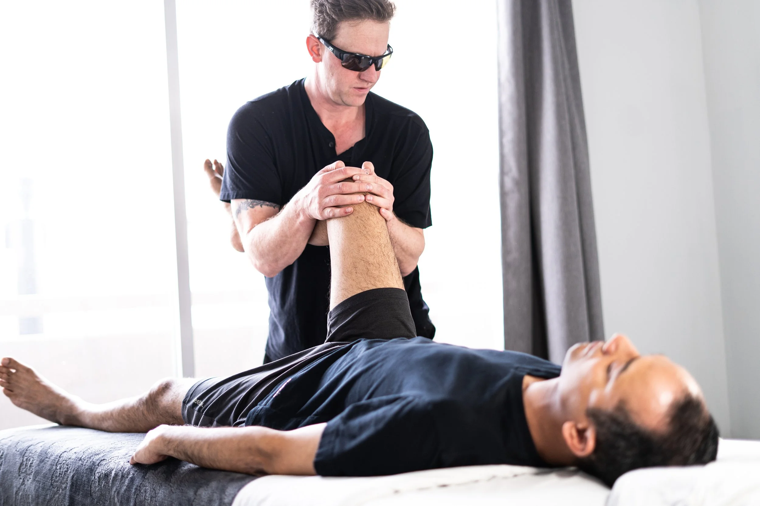 David wearing sun glasses performing a leg assessment on a client's hip and knee during therapy in a well-lit room.