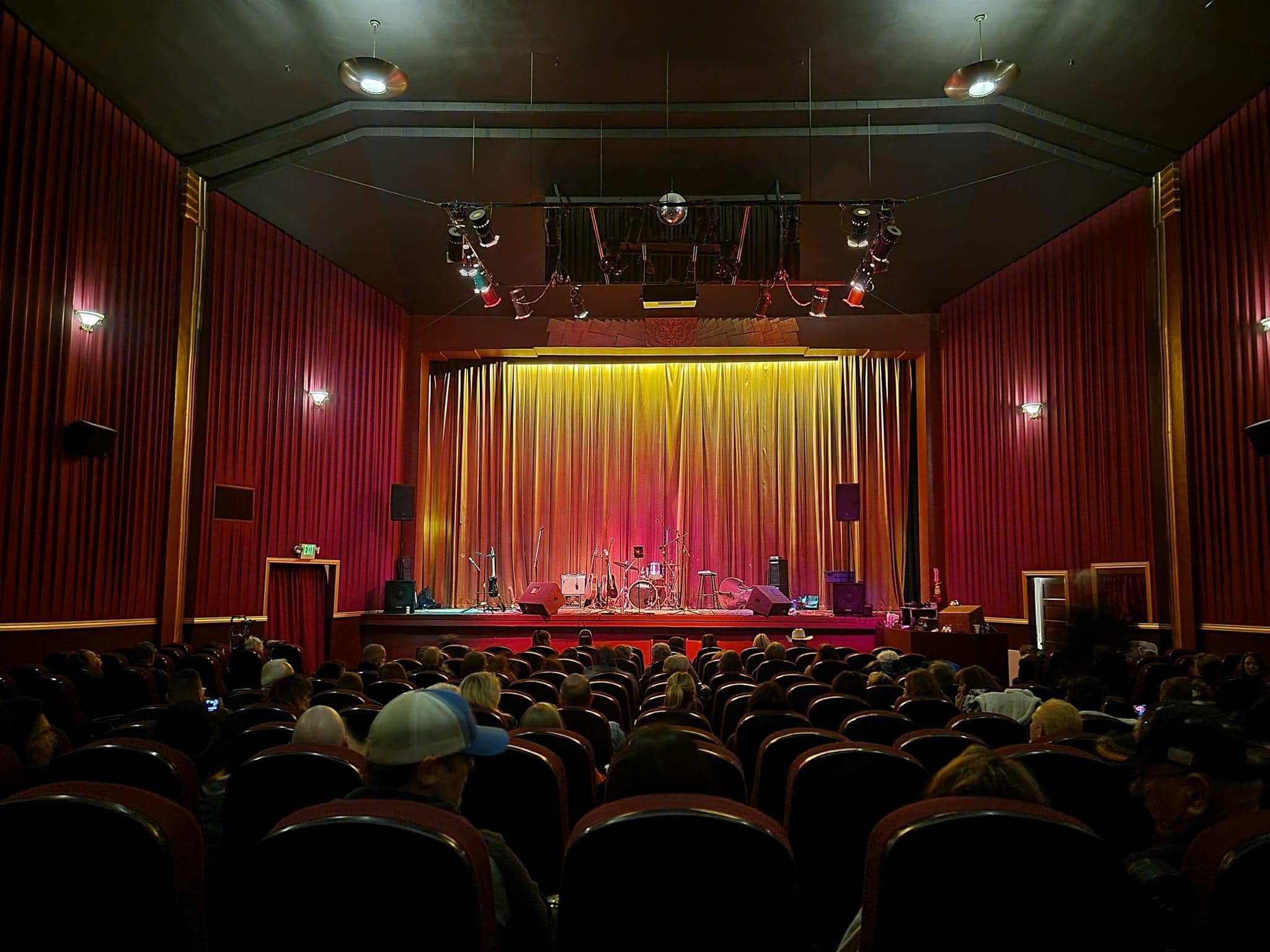 Empty stage with musical instruments and microphones in front of closed gold curtains, in a theater with red velvet walls and an audience seated facing the stage.