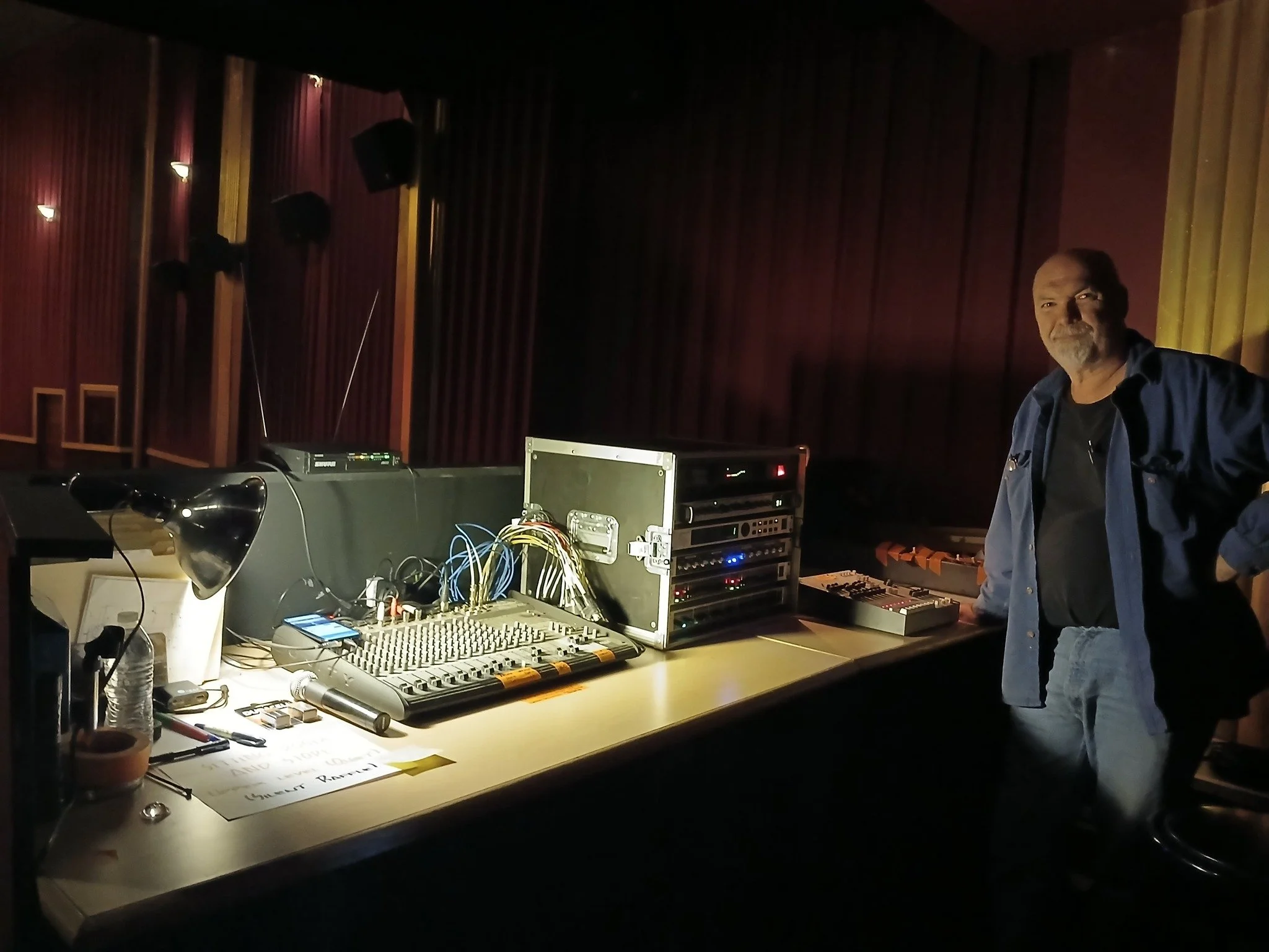 Man standing next to sound equipment and mixing console at a music or recording studio with dim lighting and red curtains.