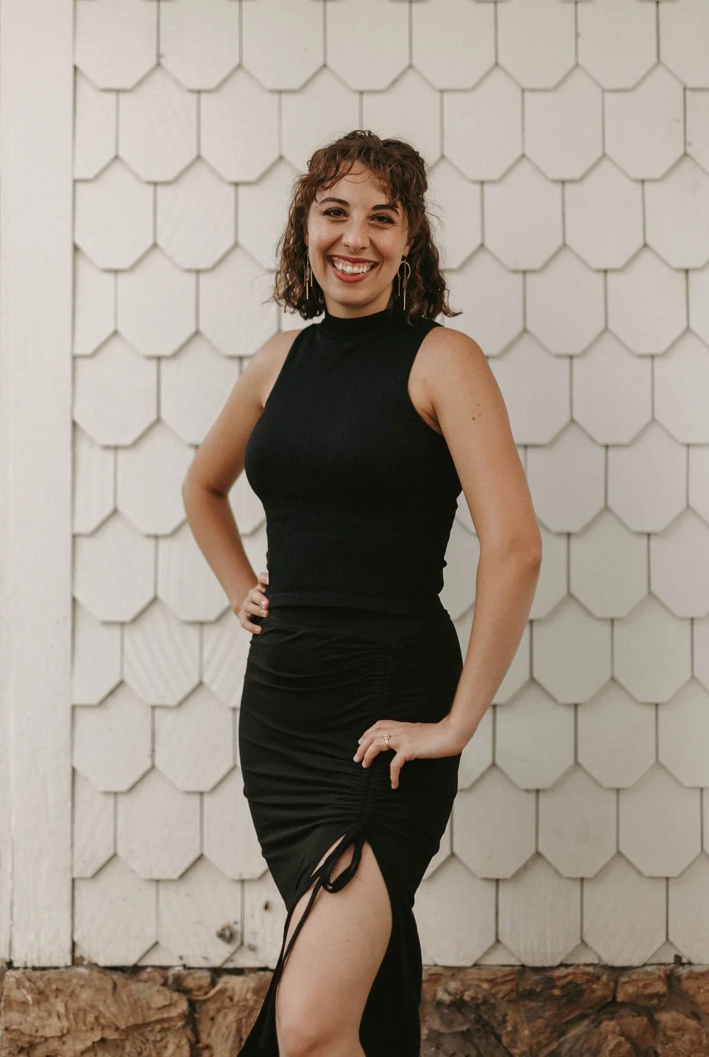 A woman with curly brown hair wearing a sleeveless black top and a black ruched skirt with a slit, standing in front of a white hexagonal tiled wall, smiling with her right hand on her hip.
