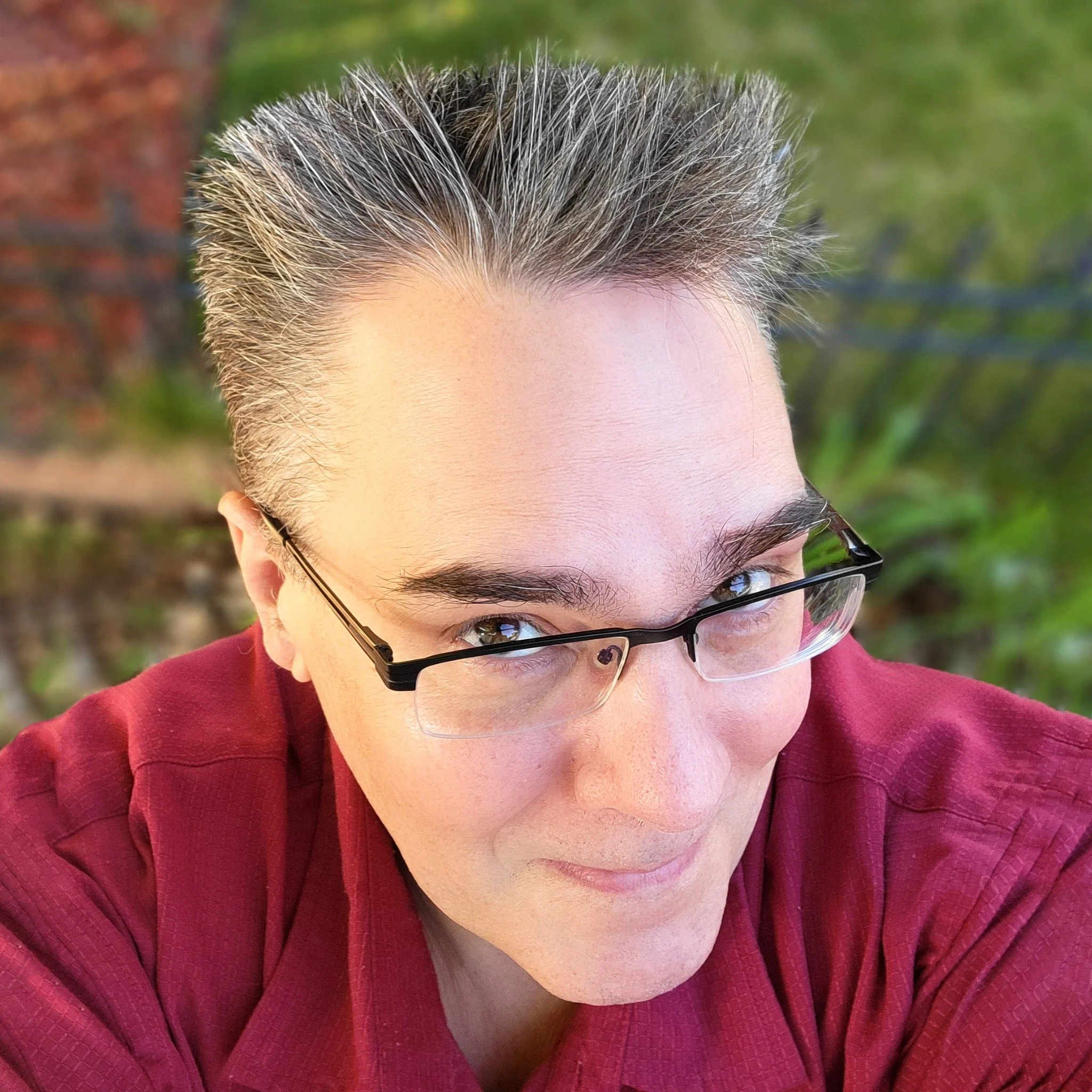 A man with glasses taking a selfie outdoors, wearing a red shirt, with a blurred background of greenery and a brick wall.