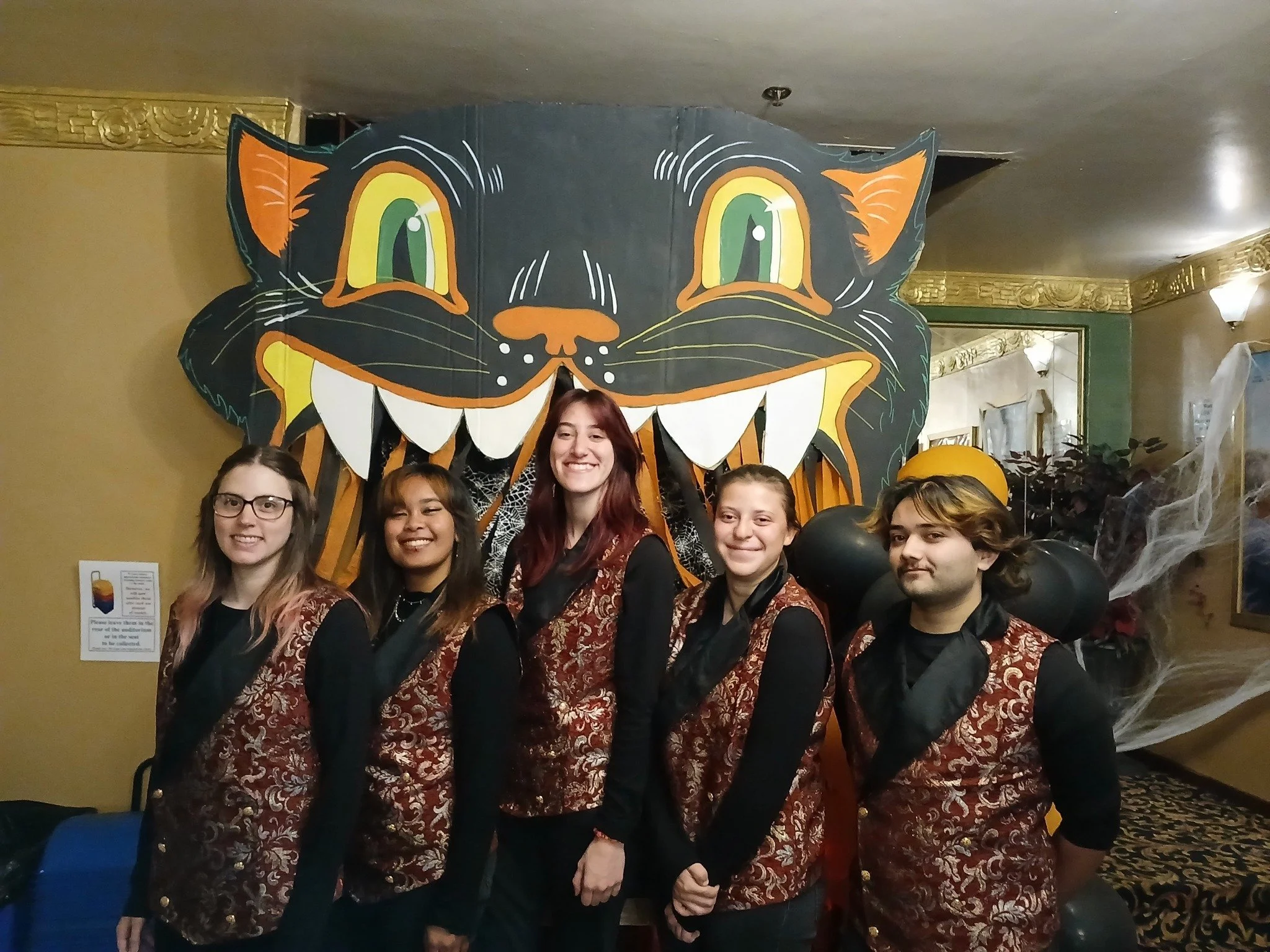 Five people standing in front of a large cartoonish black cat face Halloween decoration with large eyes and sharp teeth, dressed in matching patterned vests.