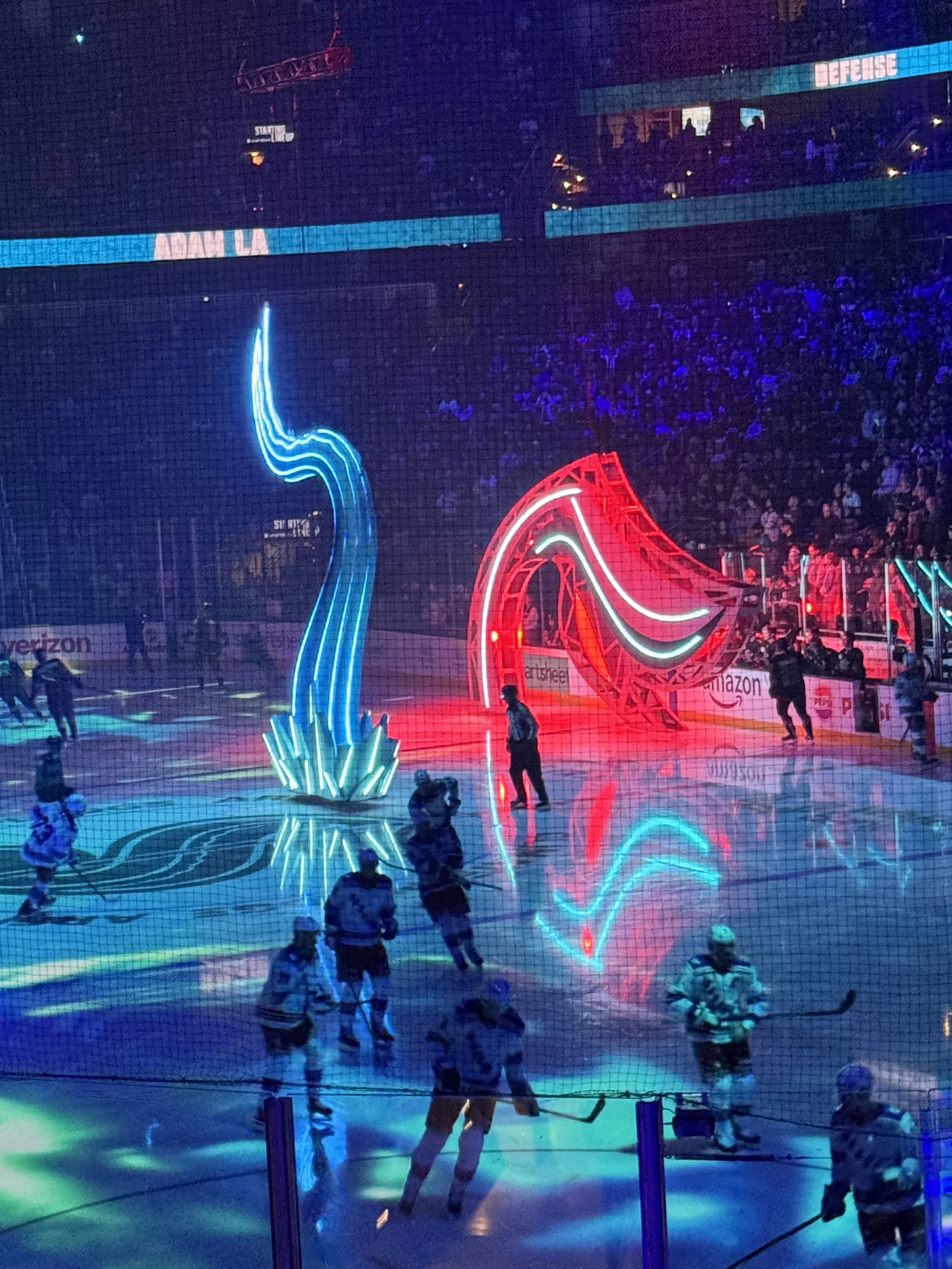 An ice hockey arena with players on the ice, vibrant neon lights in red and blue, and a large screen displaying the name 'Adam La'. The arena has an energetic atmosphere with spectators in the background and neon light installations near the ice.