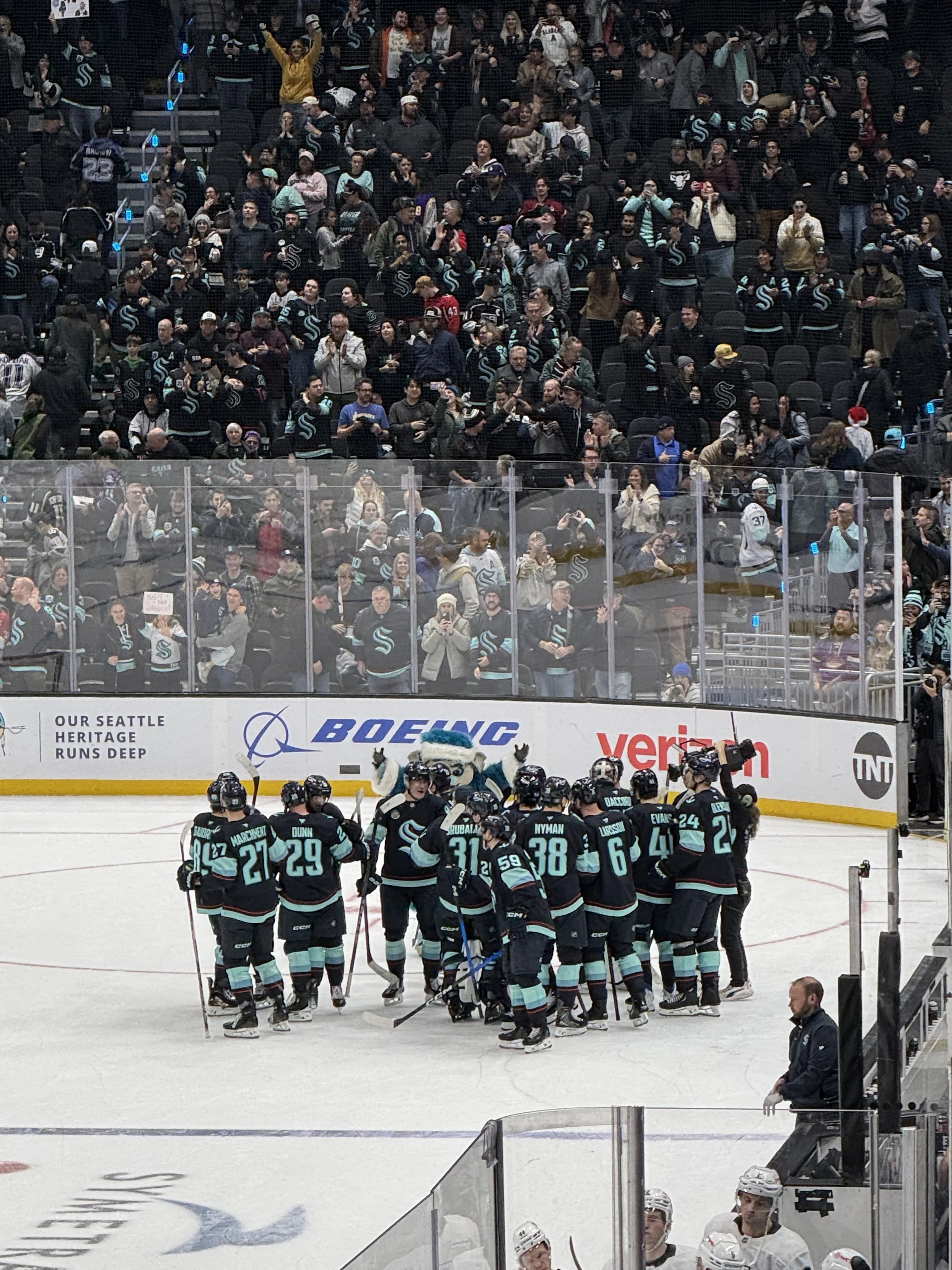 Ice hockey team in dark jerseys with teal accents celebrating on the ice at a game, with spectators in the stands cheering behind the glass.