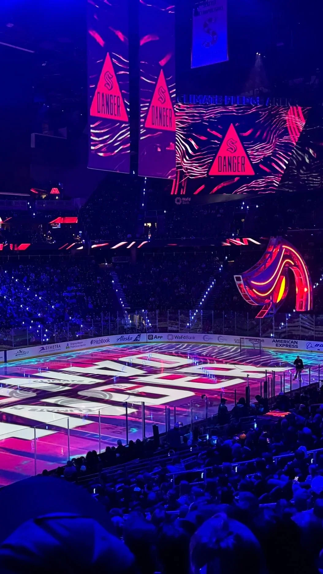 Inside an ice hockey arena with a mostly dark background, illuminated with neon pink and purple lighting. The ice rink features the Seattle Kraken logo, and there are large LED screens displaying warning signs with the word 'DANGER' and a stylized 'S' symbol.