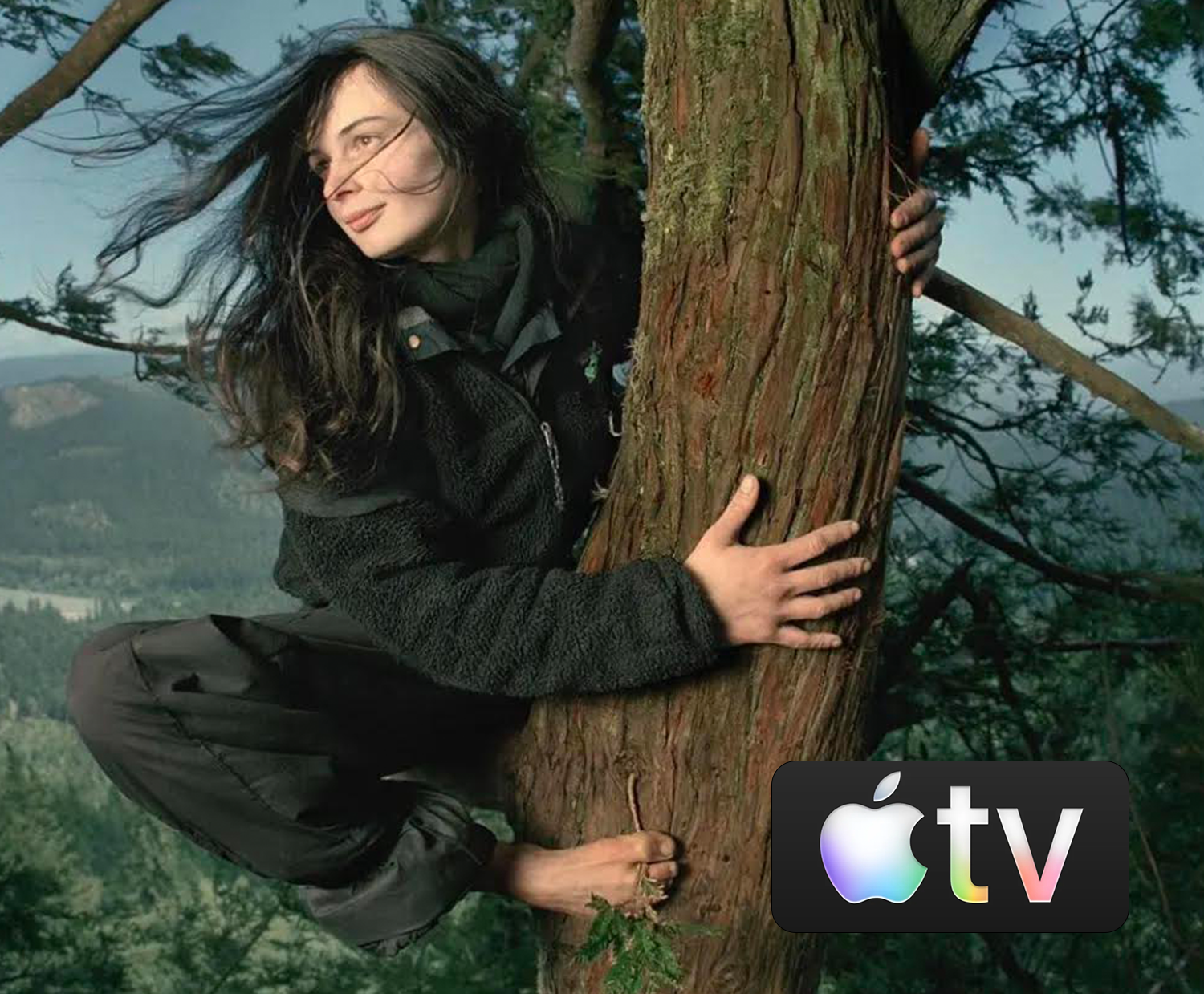 A woman with long dark hair, wearing a black jacket, hugging a tree in a forested area during daytime, with mountains and trees in the background.