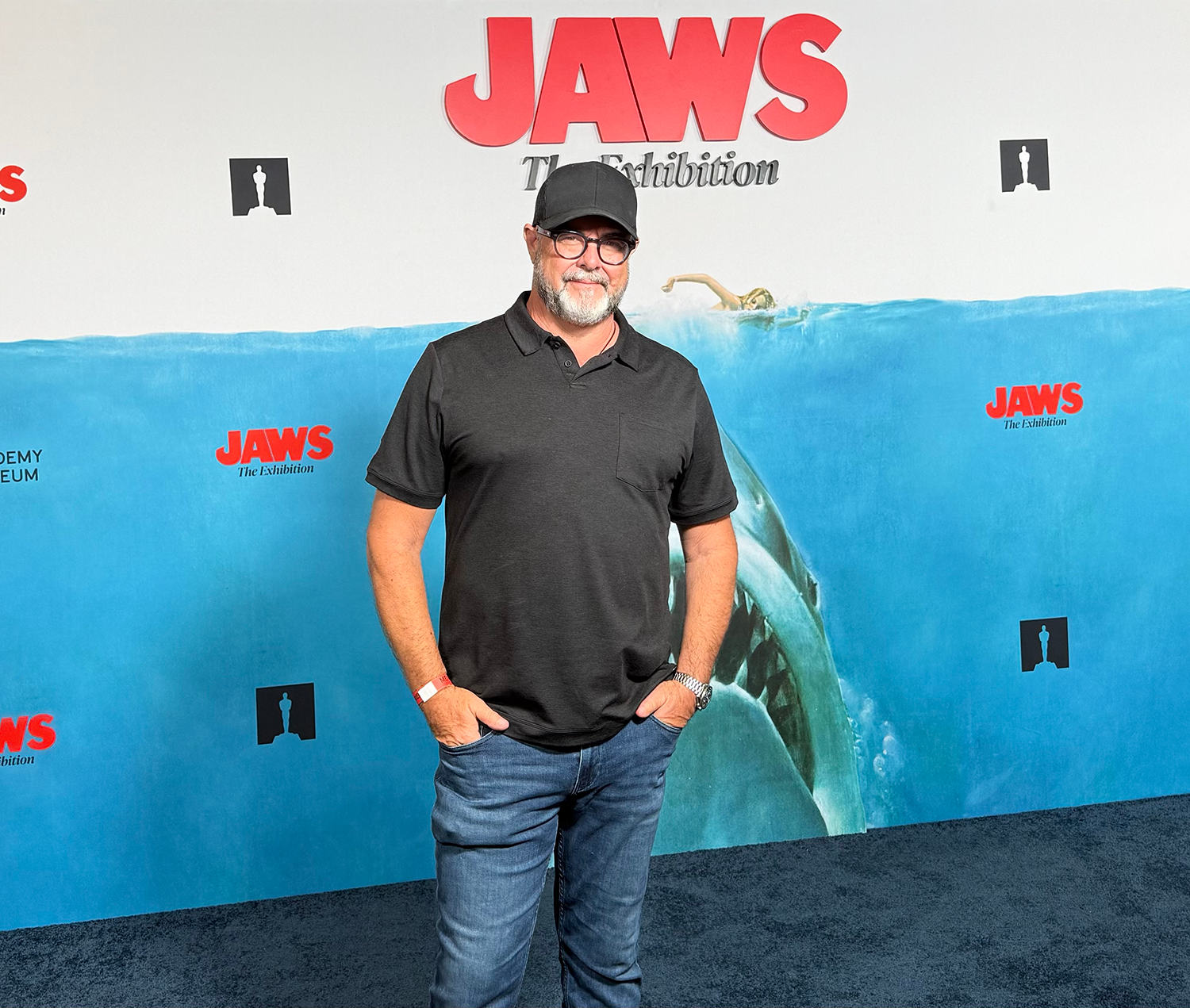 A man with glasses, a beard, and a black cap standing in front of a JAWS exhibition backdrop, wearing a black polo shirt and jeans.