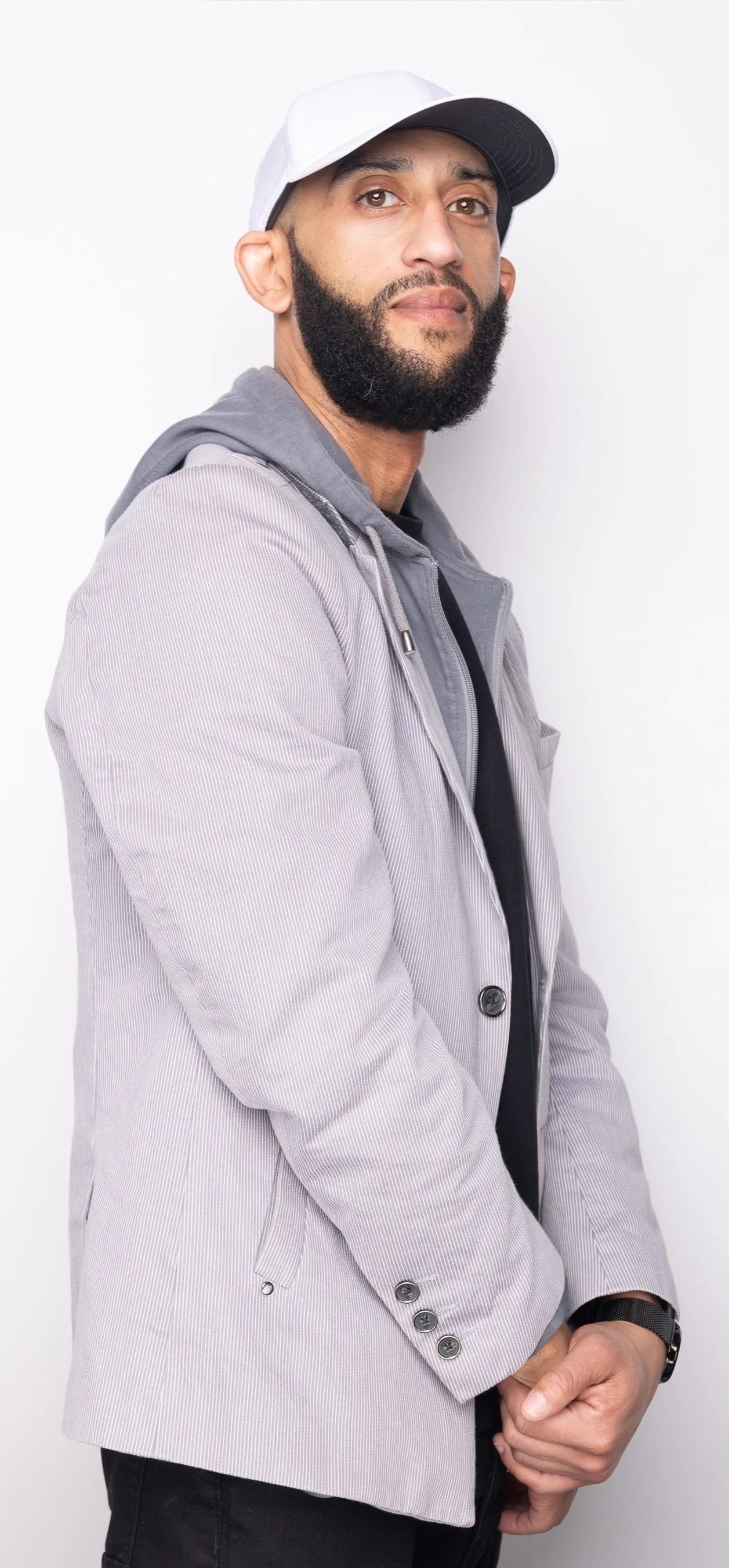 A man with a beard wearing a white cap, gray jacket, black shirt, and black pants, standing against a plain white background.