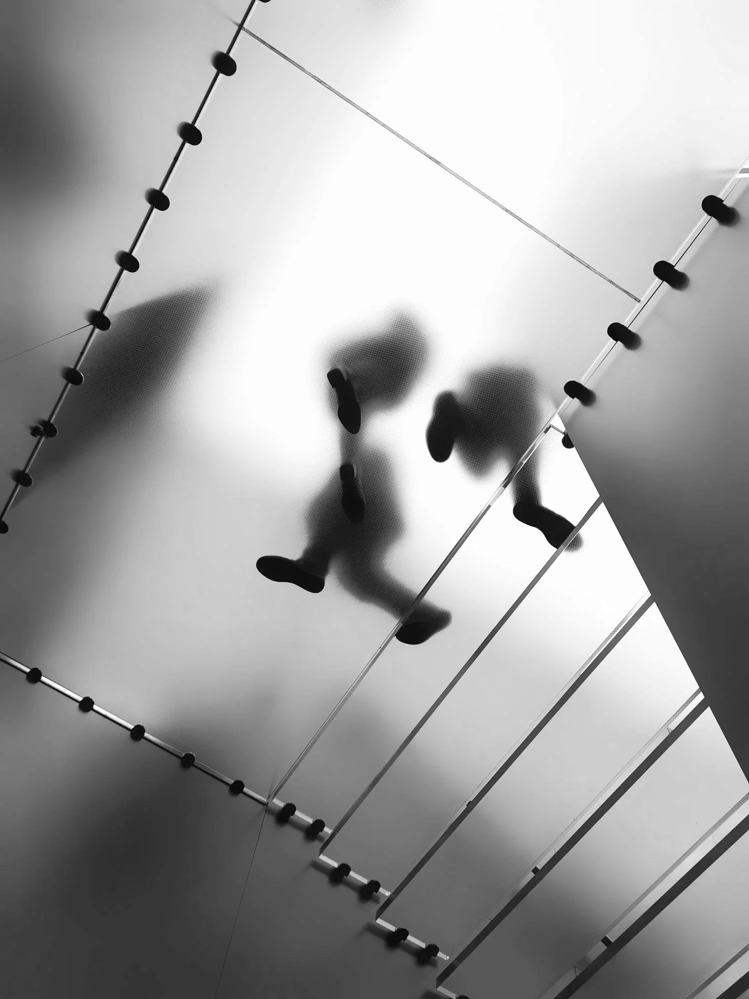 A person walking on a reflective surface with bright lighting, seen from below through a glass ceiling with black fixtures and metal bars.