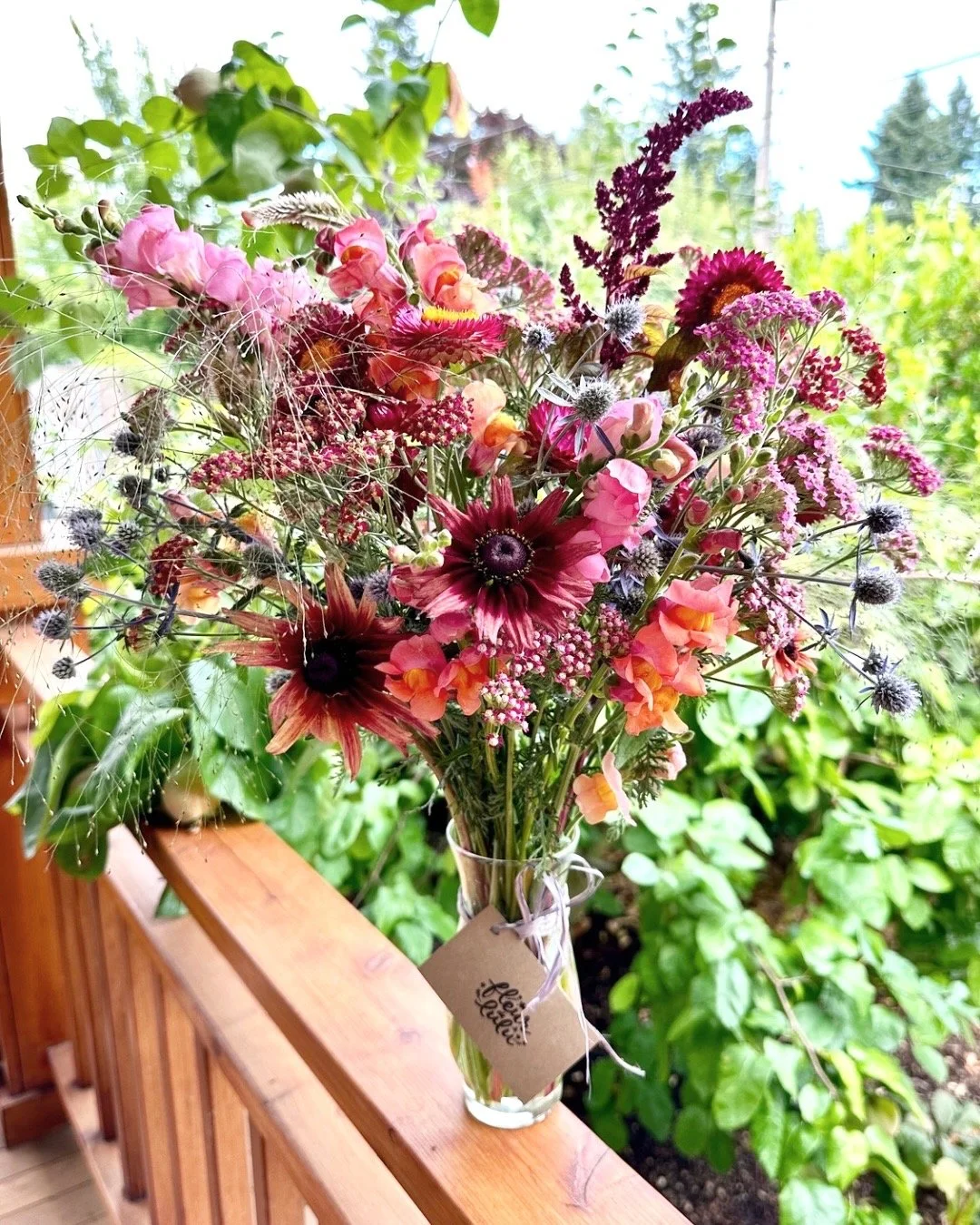 A colorful bouquet of mixed flowers in a tall glass vase on a wooden balcony railing, with greenery and trees in the background.
