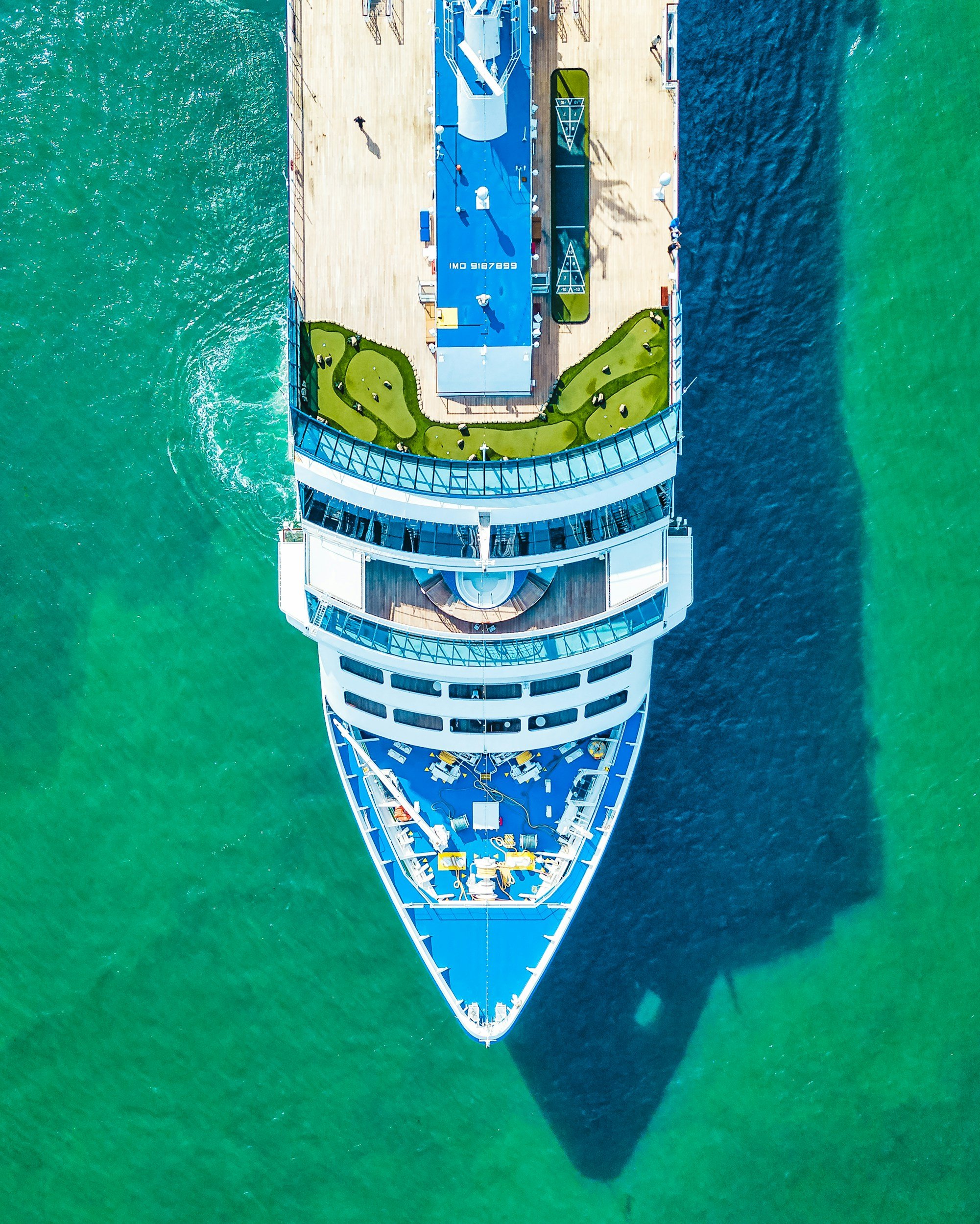 Aerial view of a large cruise ship sailing on green water, showing the top deck with a mini golf course, outdoor seating, and various amenities.