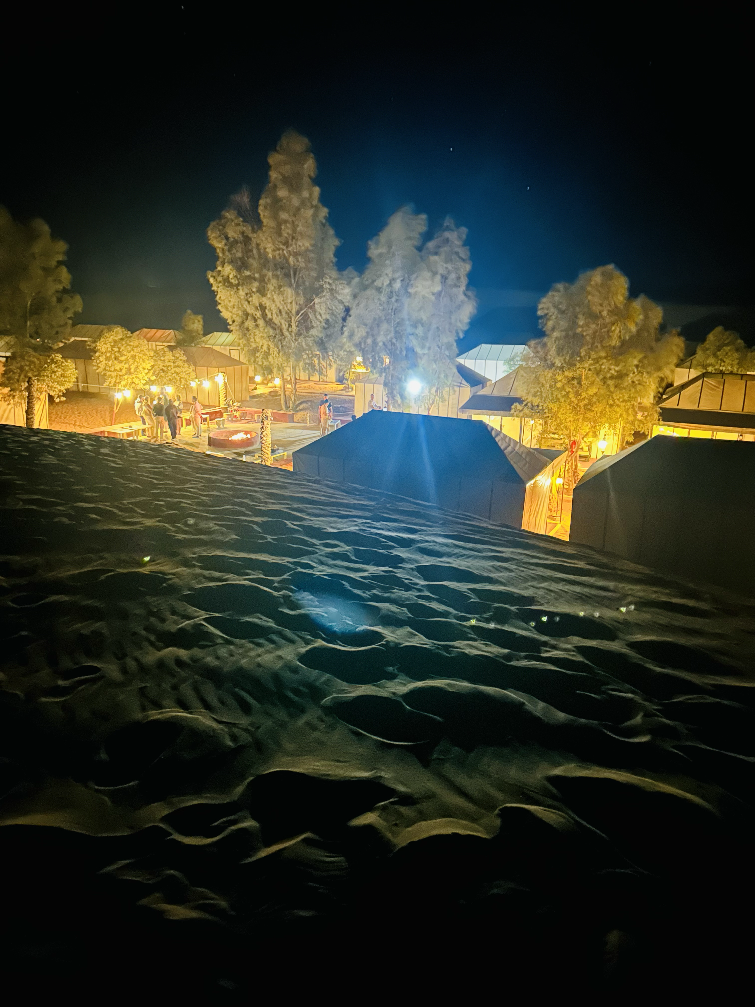Nighttime scene at a beach or desert with sand dunes in the foreground, illuminated tents and trees in the background, and a dark sky with a few stars.