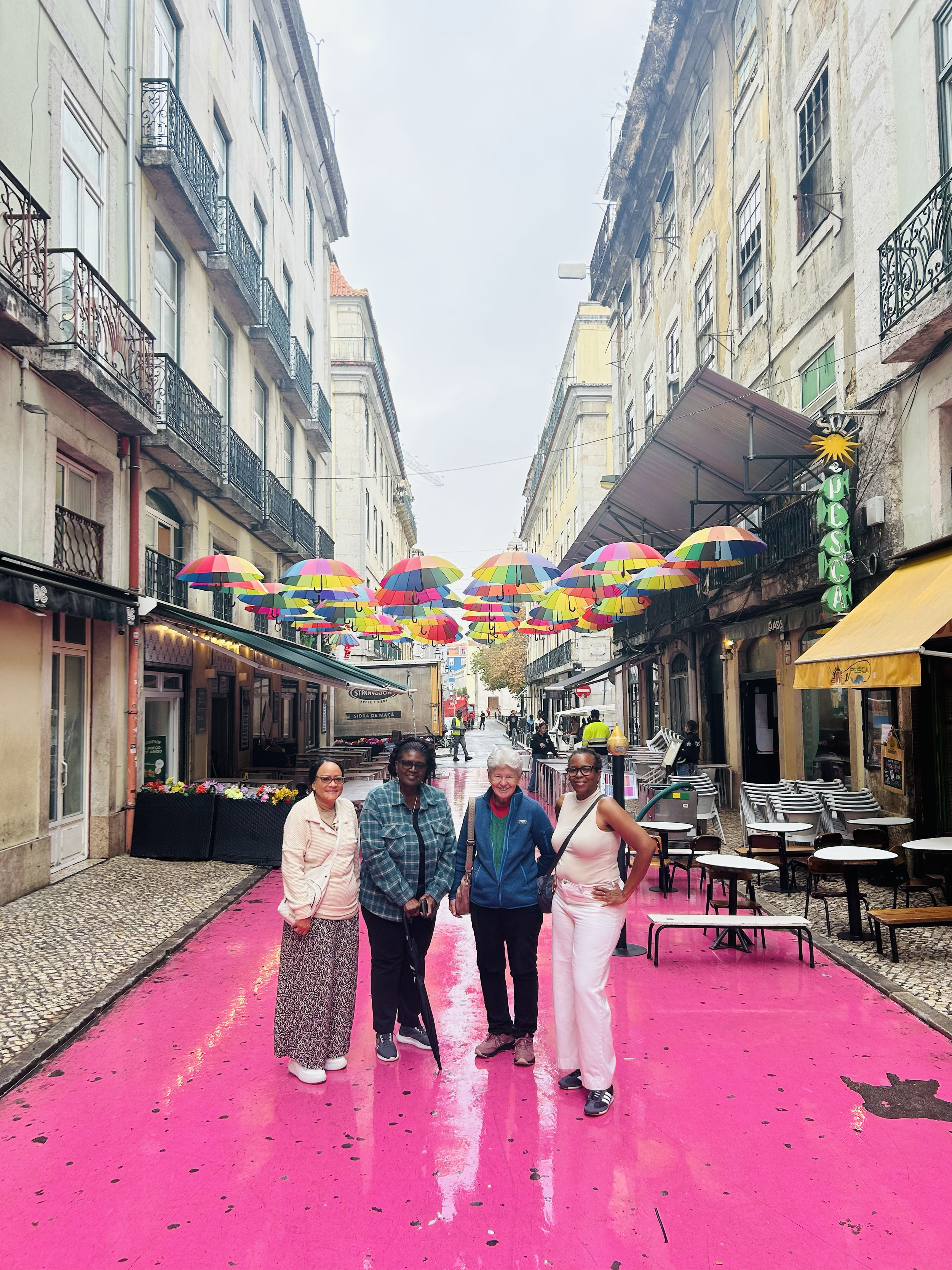 Four women standing on a pink street in a European city, with colorful umbrellas hanging overhead and outdoor cafes visible on the sides.