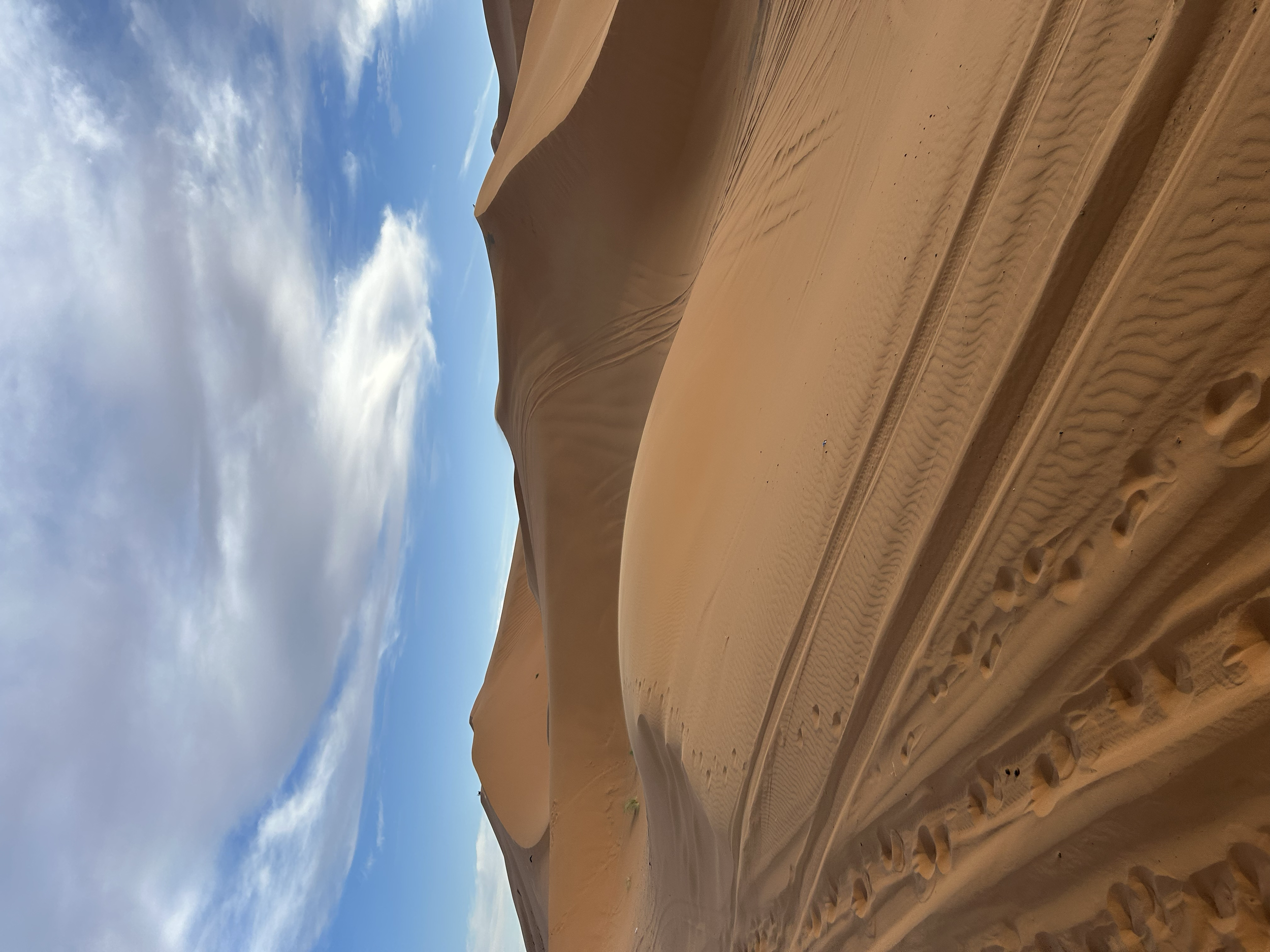 Dunes in a desert with tire tracks in the sand and a partly cloudy sky above.