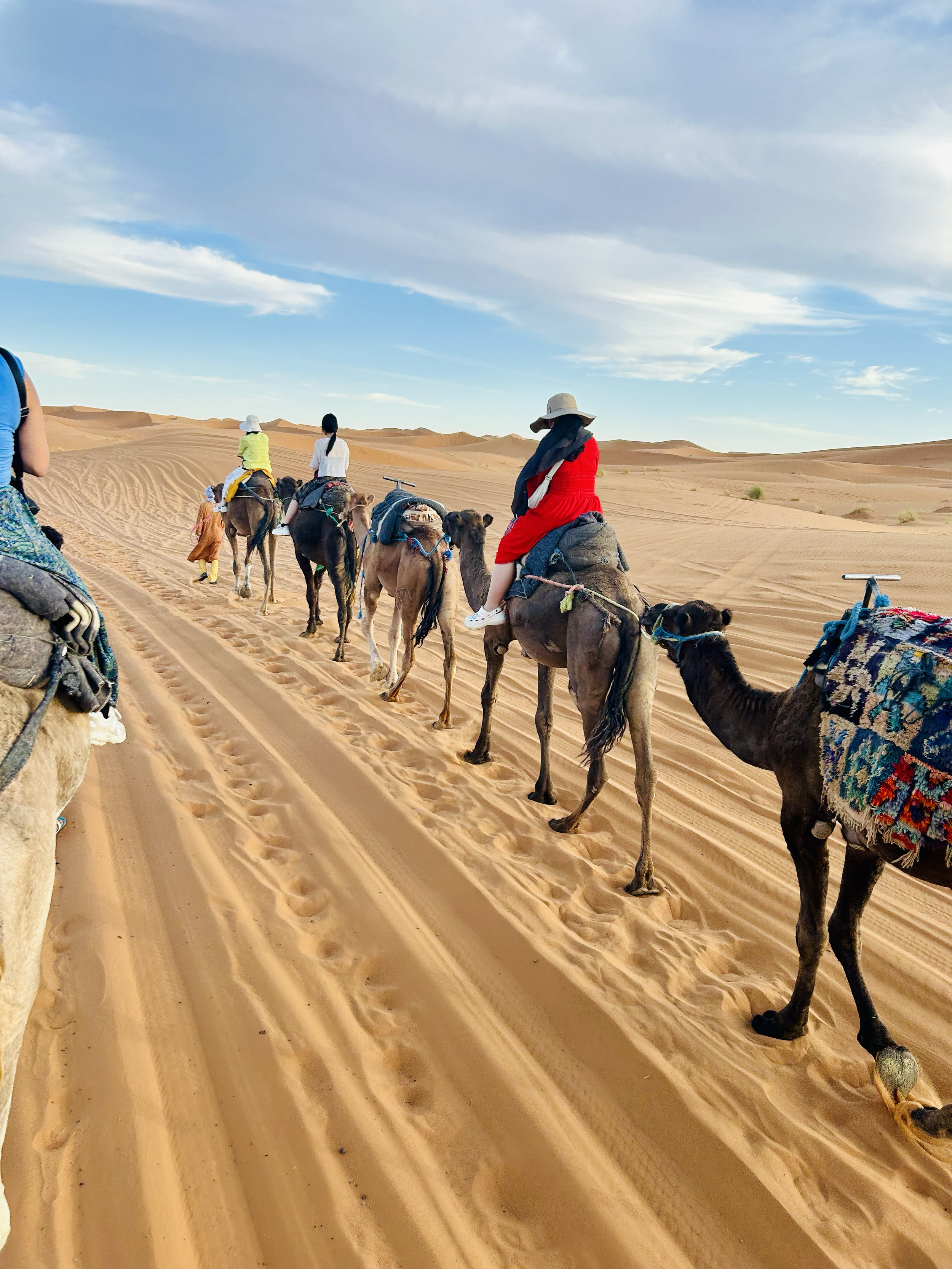 A group of people riding camels through a desert landscape with sand dunes under a partly cloudy sky.
