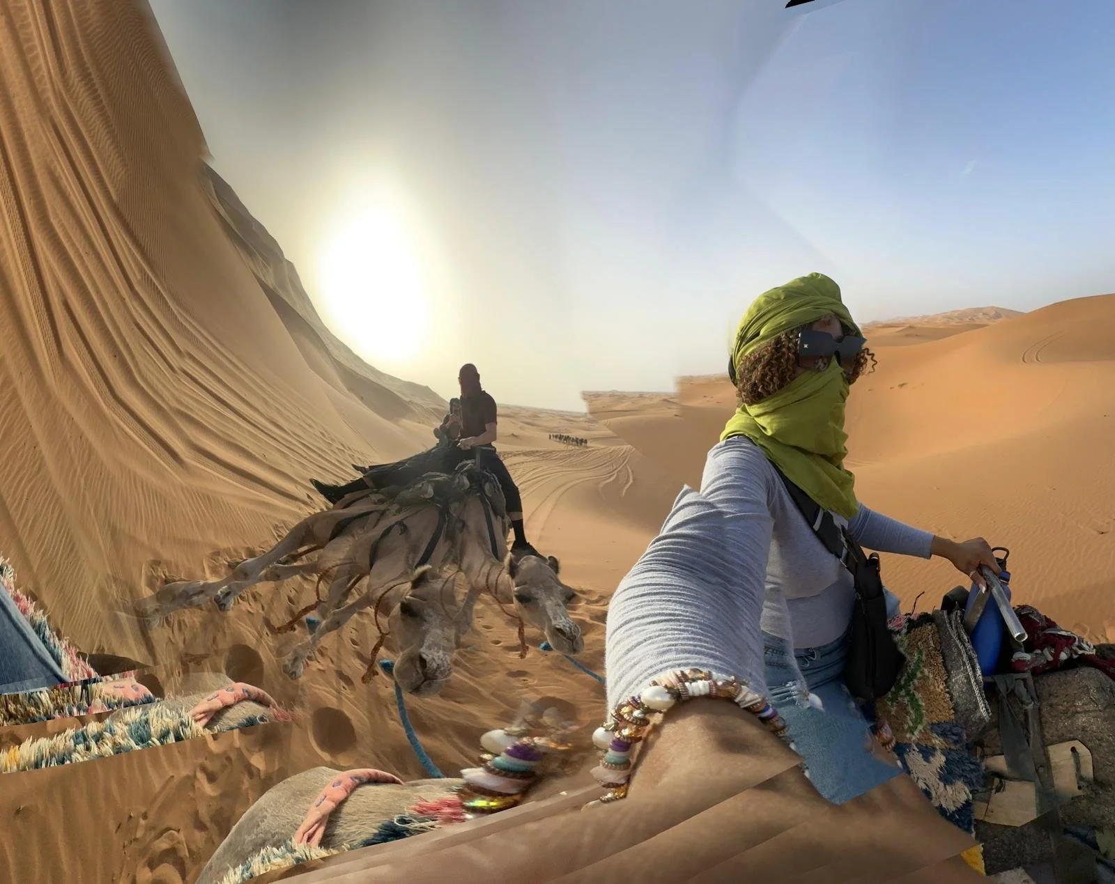 A person in desert attire, wearing sunglasses, a green headscarf, and bracelets, taking a selfie while riding a camel in a desert landscape with sand dunes and a clear blue sky.