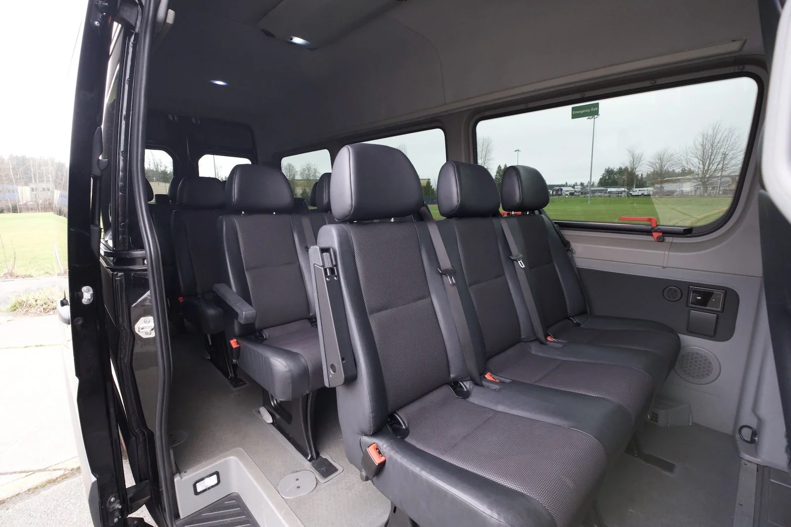 Interior of a passenger van with three rows of black fabric seats, seat belts, large side windows, and an emergency exit sign outside.