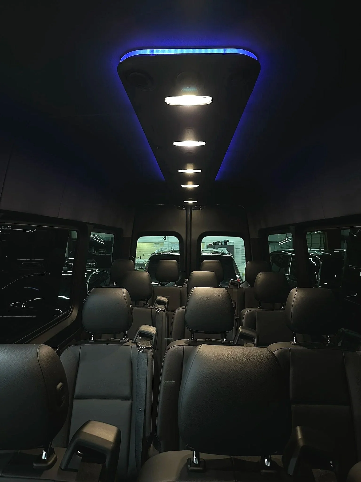 Inside view of a vehicle with multiple rows of gray seats, overhead ceiling lights, and windows showing parked cars outside.