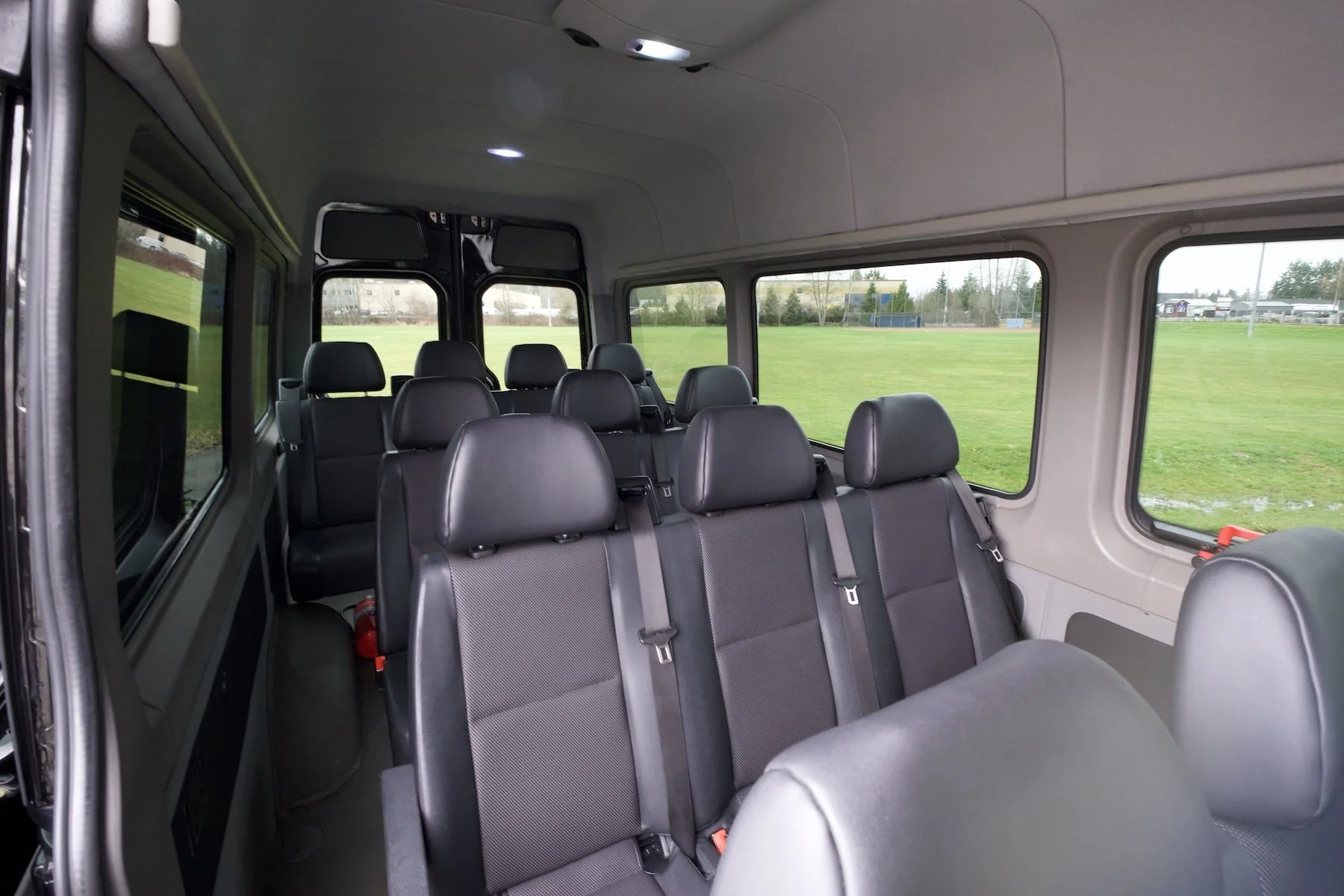 Interior of a passenger van with multiple black seats, large windows, and a view of green grass outside.