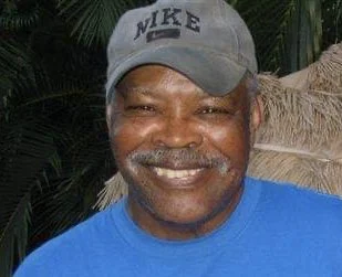 A smiling man wearing a gray Nike baseball cap and a blue shirt, with palm leaves in the background.