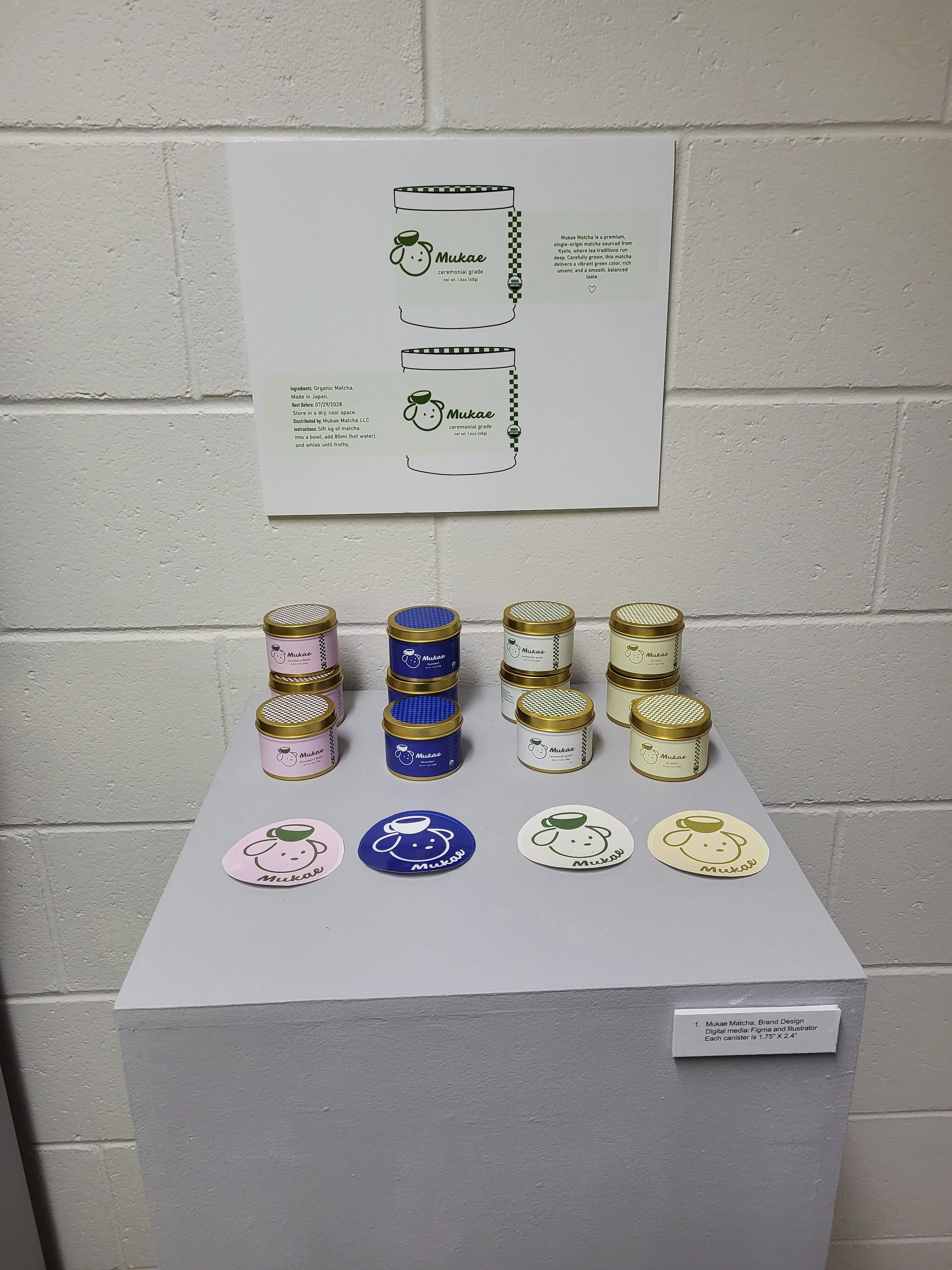 Display of small matcha tea tins in pink, blue, white, and yellow on a white table, with corresponding stickers and an informational poster on a white brick wall behind.