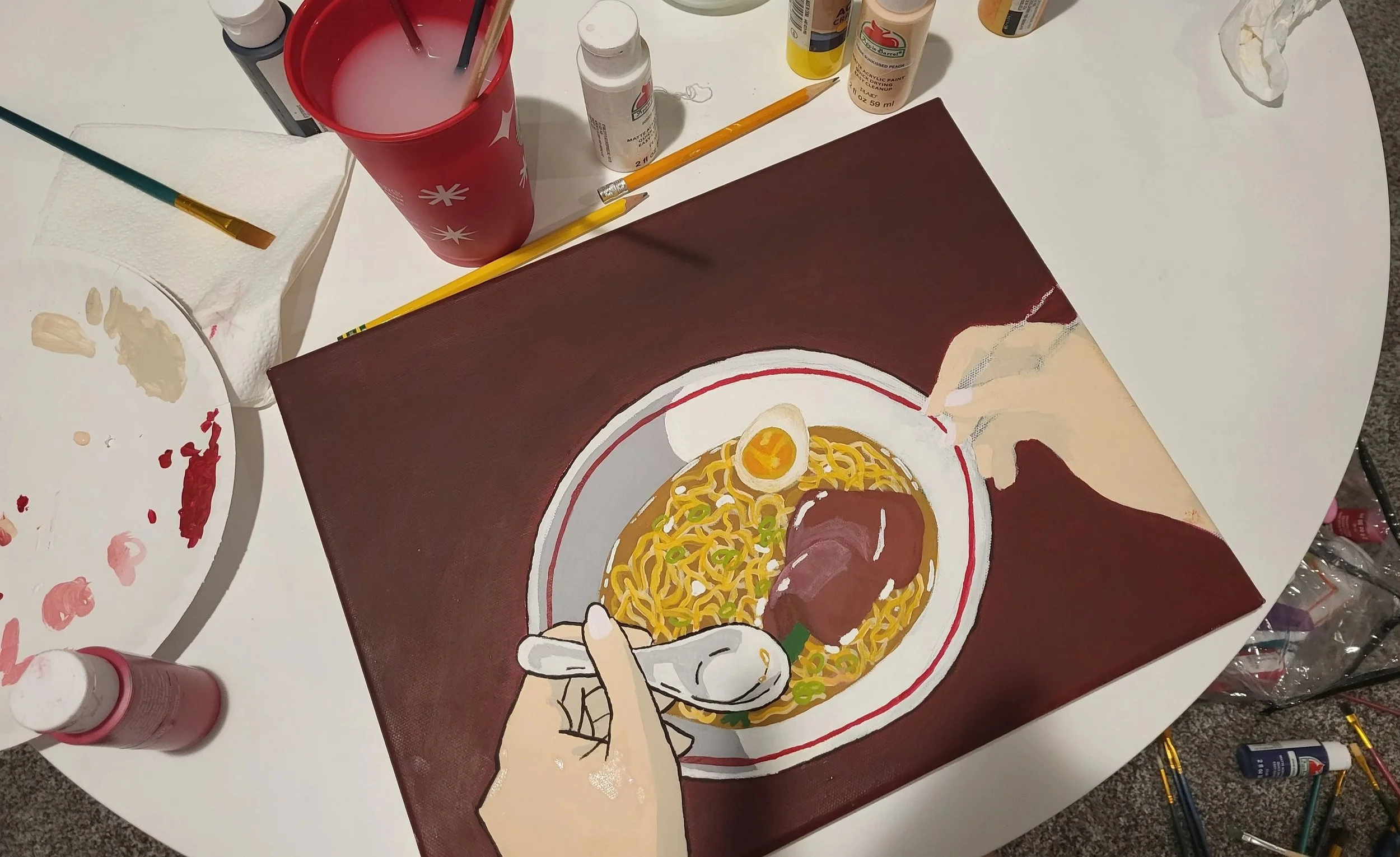 A painting of a bowl of ramen with an egg, chopsticks, and green onions, on a brown background, surrounded by painting supplies, including brushes, paint bottles, a paper towel, and a palette.