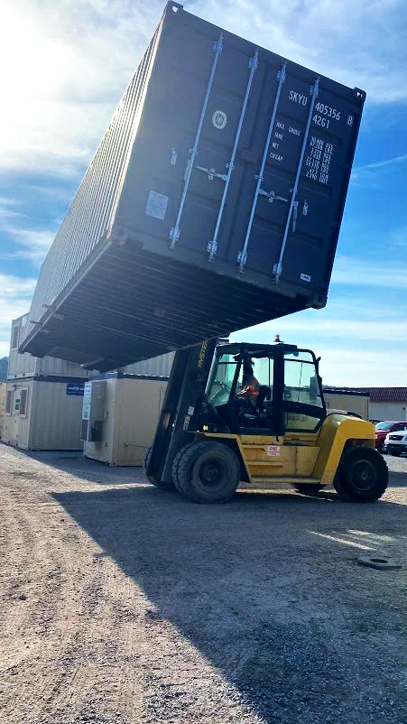 A forklift lifting a large shipping container in an outdoor yard with partly cloudy skies.