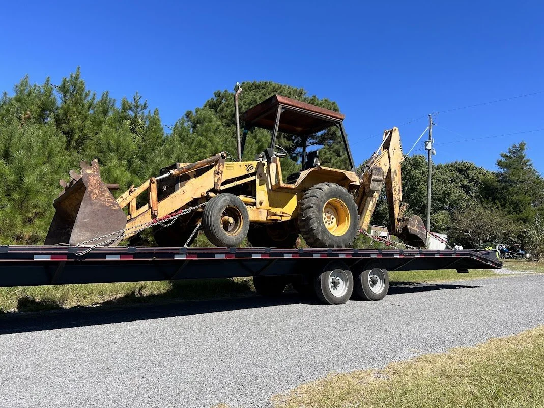 Yellow bulldozer being transported on a flatbed trailer on the side of a road with trees and utility poles in the background.