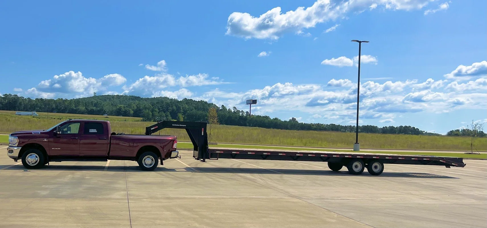 A maroon pickup truck attached to a flatbed trailer parked in an empty lot under a partly cloudy sky with green hills and trees in the background.