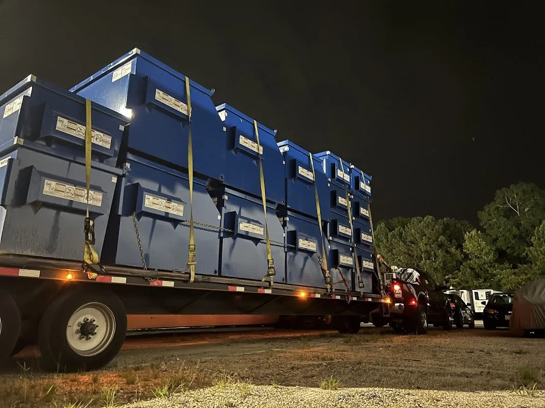 Large flatbed truck carrying blue plastic bins secured with yellow straps at night, parked alongside several other vehicles.