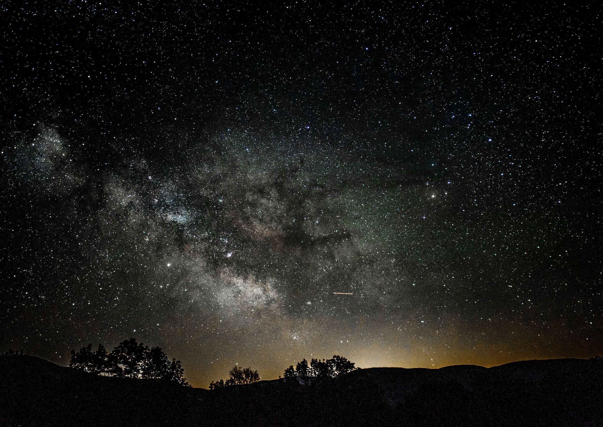 Star-filled night sky with the Milky Way galaxy, silhouettes of trees and hills on the horizon.