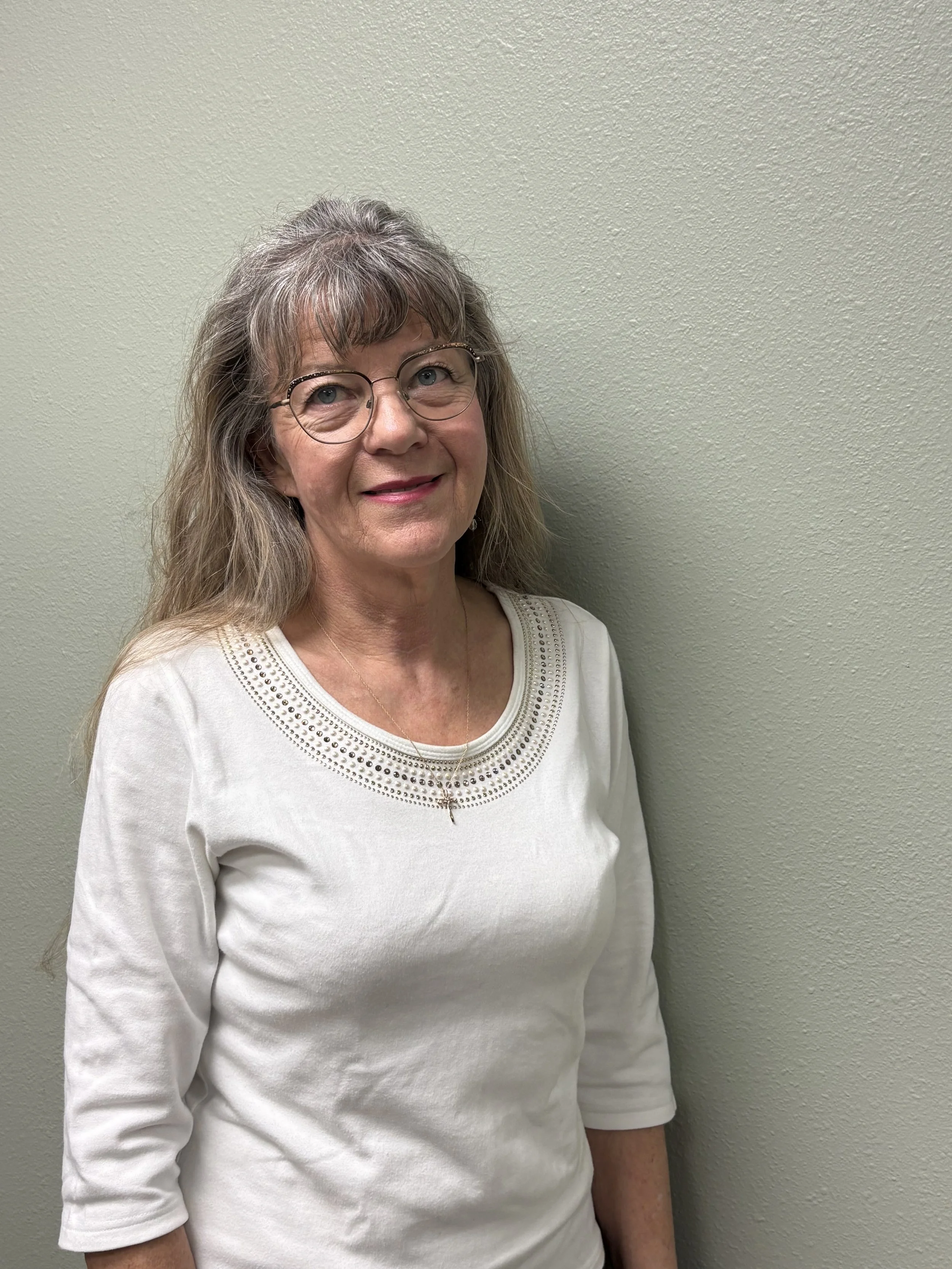 A woman with glasses and long gray hair standing against a beige wall, wearing a white top with decorative stitching around the neckline.