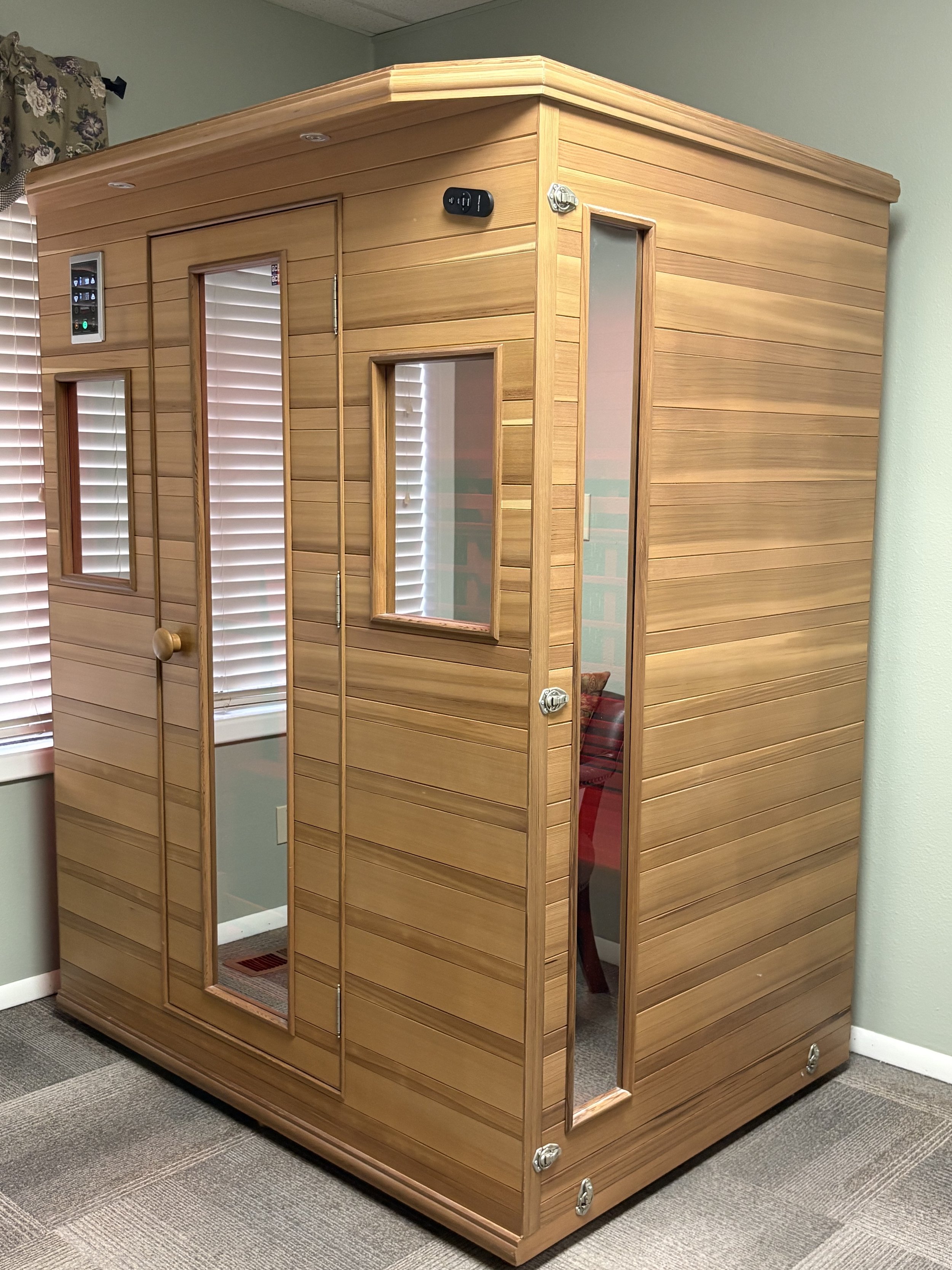 A wooden infrared sauna with glass windows and a door, placed in a room with beige carpet and light green walls.