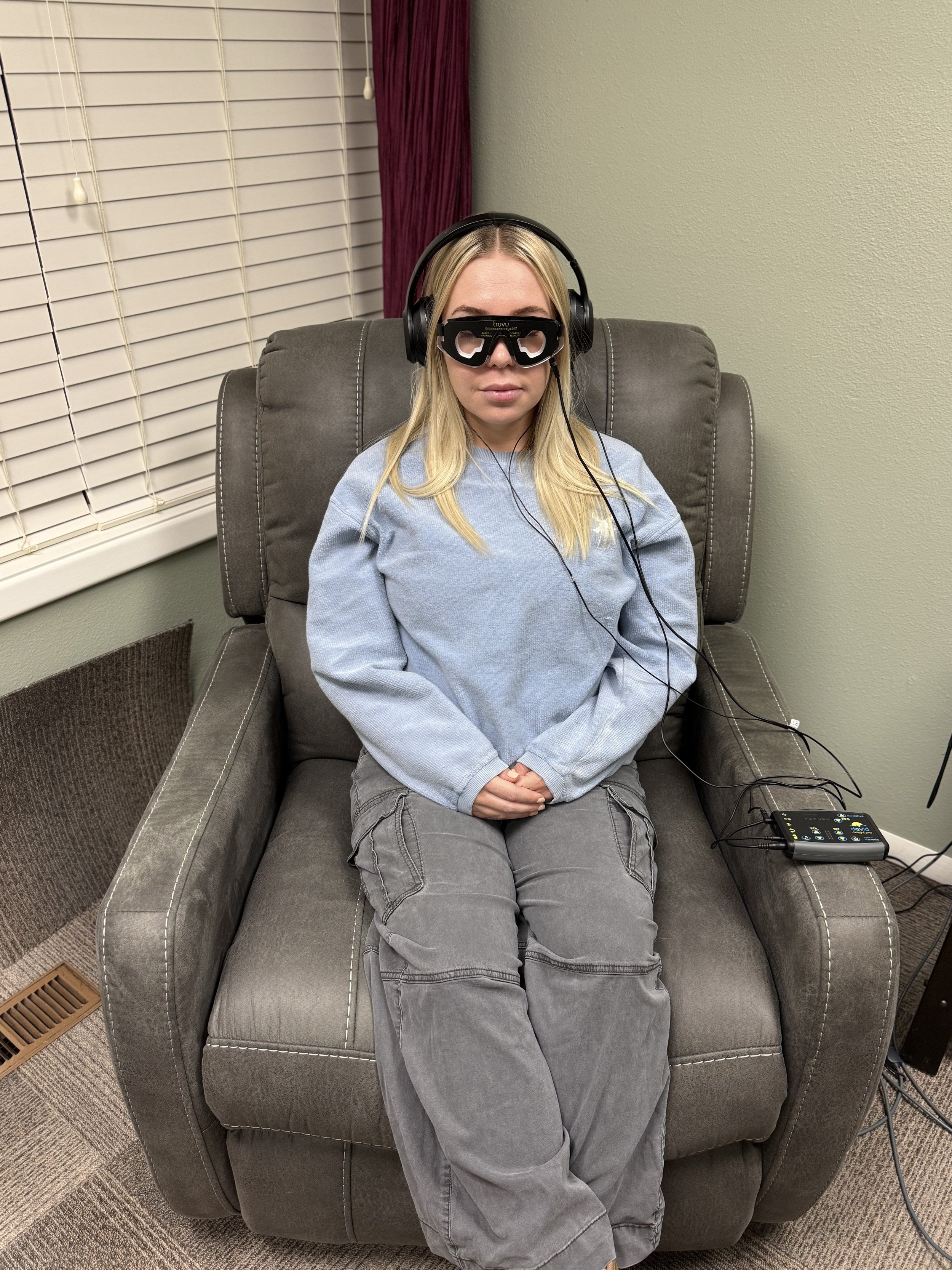 Woman sitting in a recliner chair wearing an eye-tracking device, headphones, and casual clothing with a window and blinds in the background.