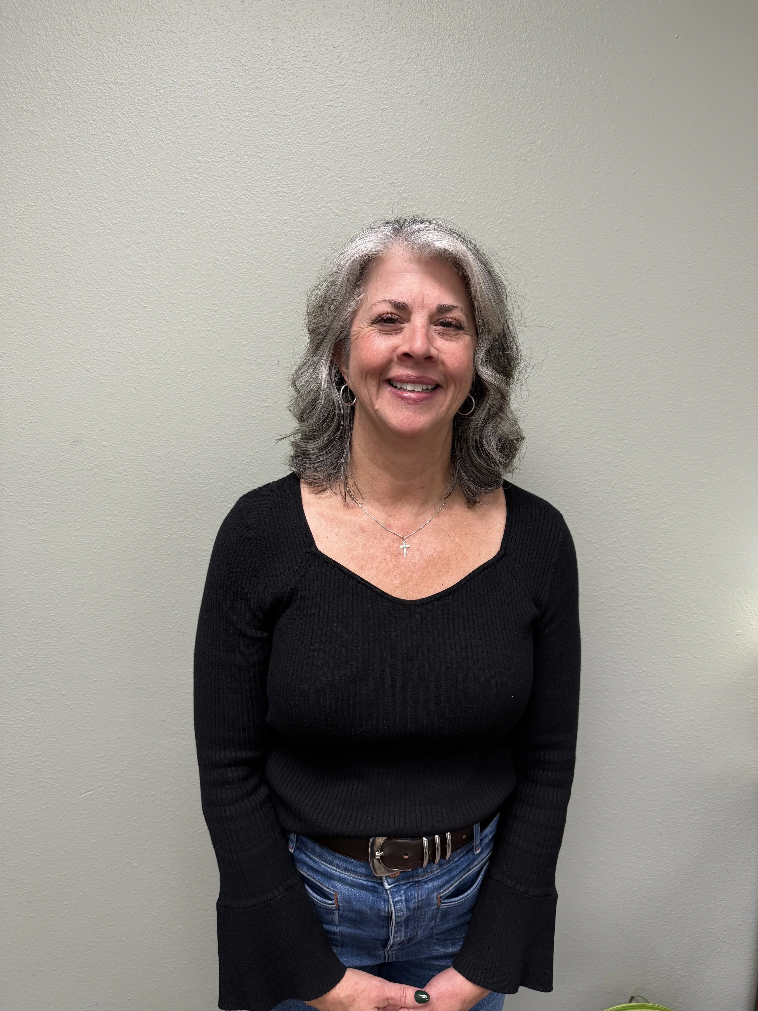 A middle-aged woman with shoulder-length gray hair, smiling, wearing a black long-sleeve sweater and blue jeans, standing against a plain light-colored wall.