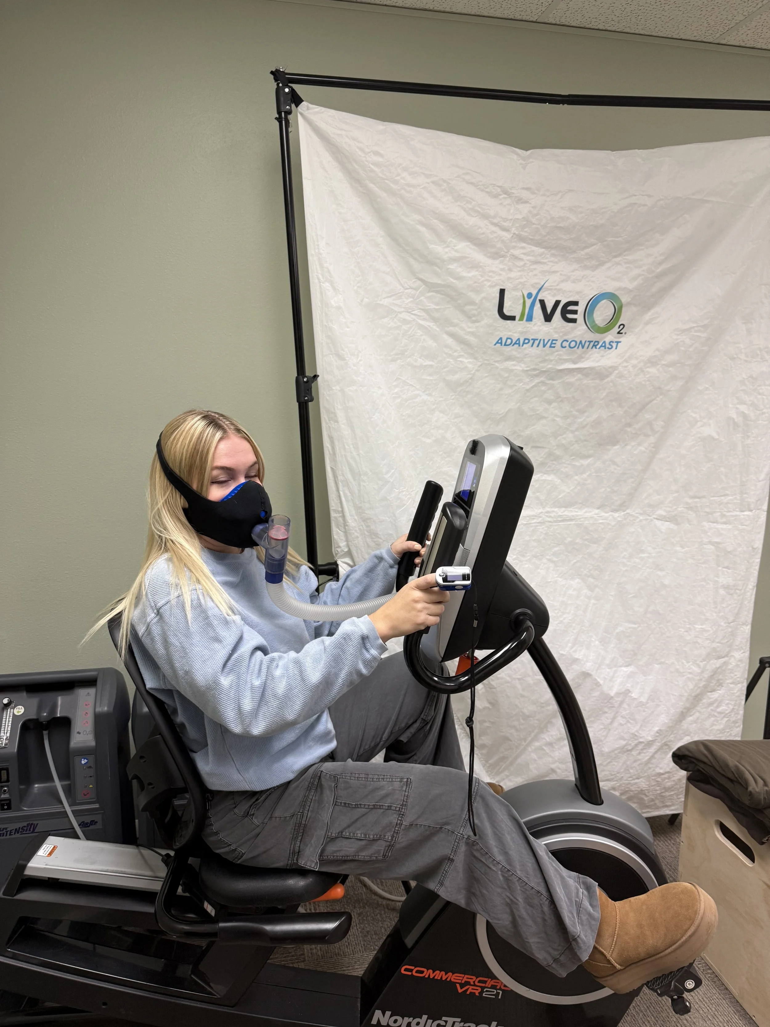 A woman using a stationary exercise bike while receiving respiratory therapy with a face mask connected to a tube, in a room with a white backdrop that says 'LIVE Q2 Adaptive Contrast'.