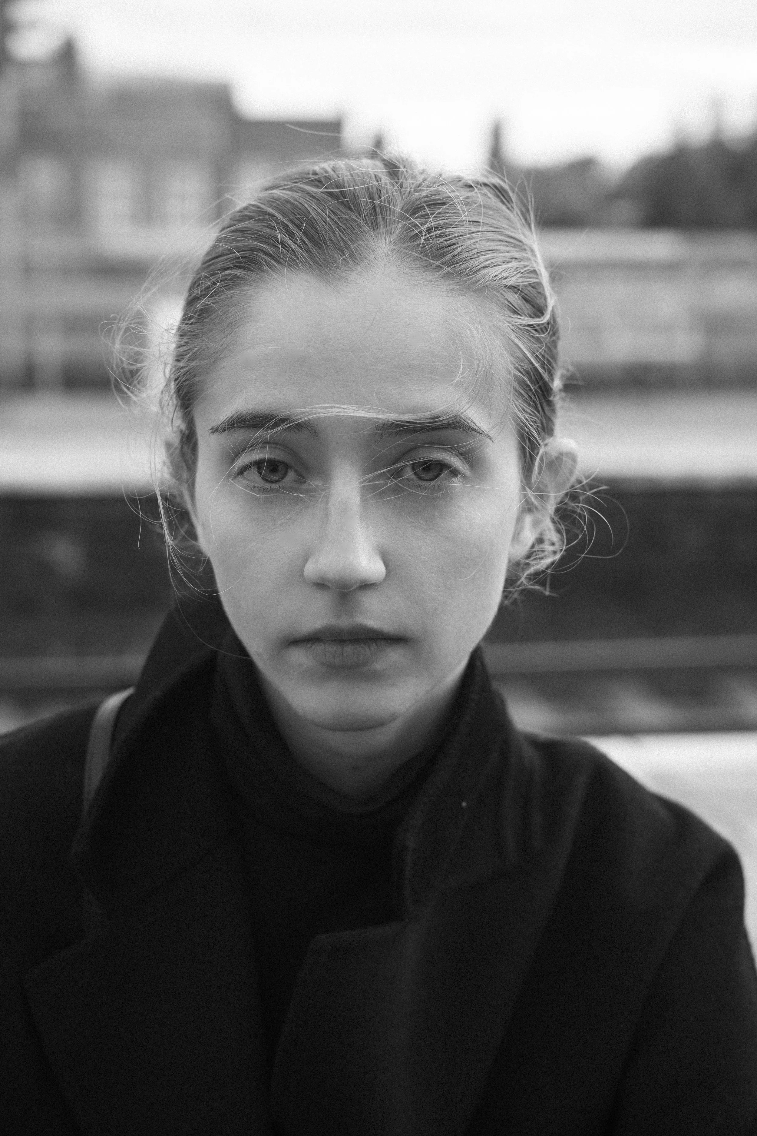 Black and white portrait of a young woman with light hair tied back, wearing a dark coat and turtleneck, looking directly at the camera with a neutral expression, outdoor background with blurred buildings.