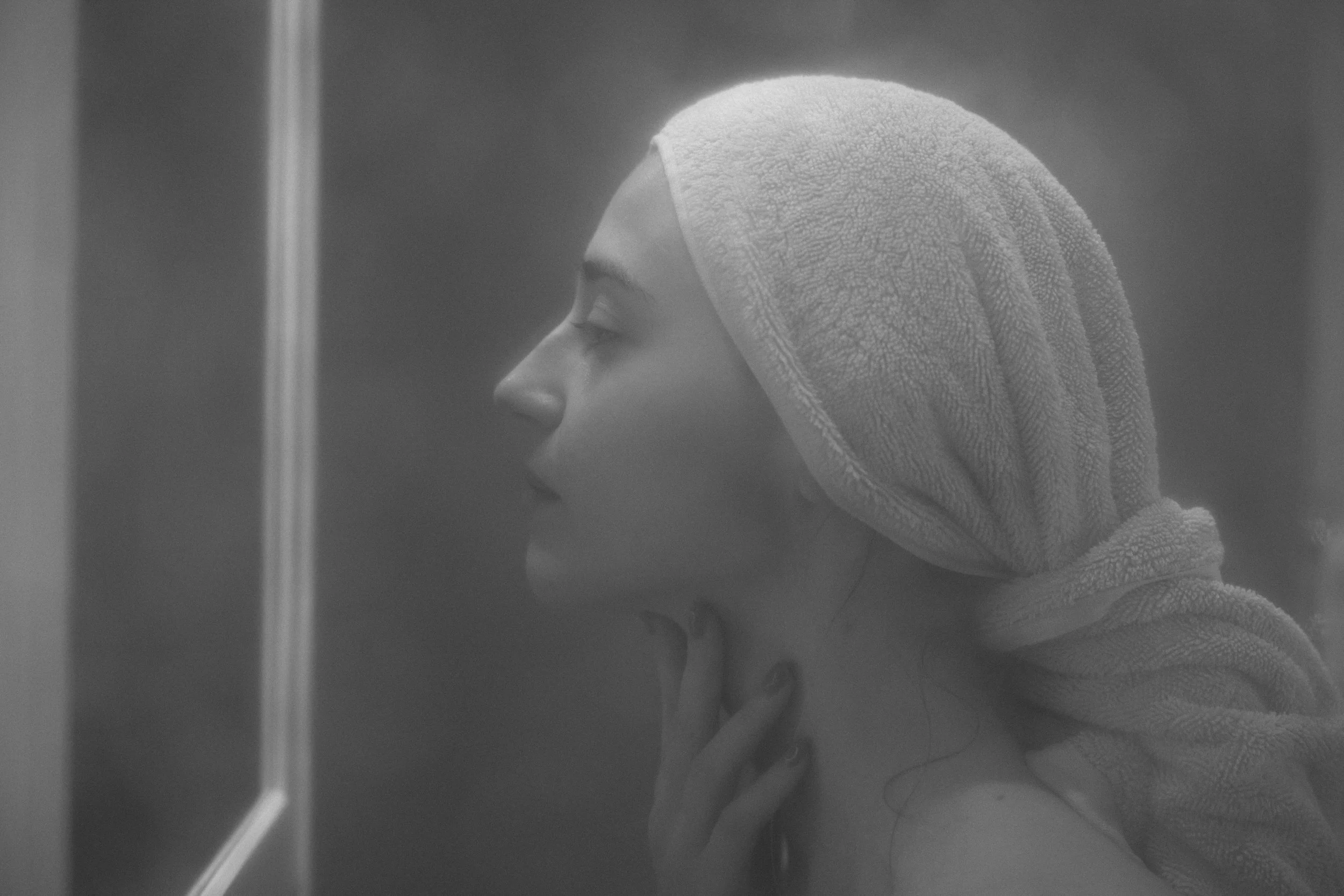 A person with a towel wrapped around their head, facing to the left, in black and white, indoors.