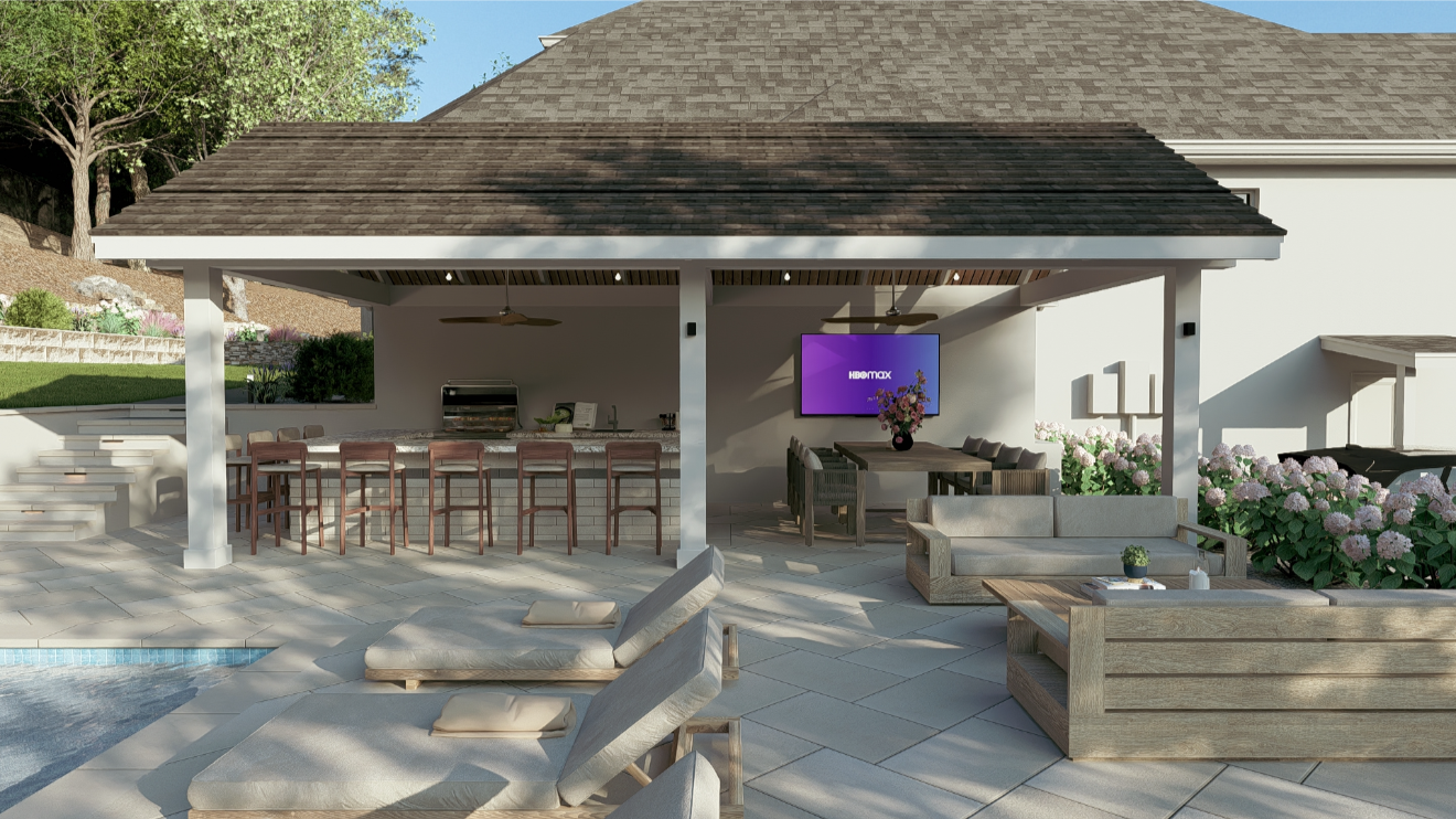 Outdoor patio area with a bar, lounge seating, and a flat-screen TV, near a house with a sloped roof and landscaped garden.