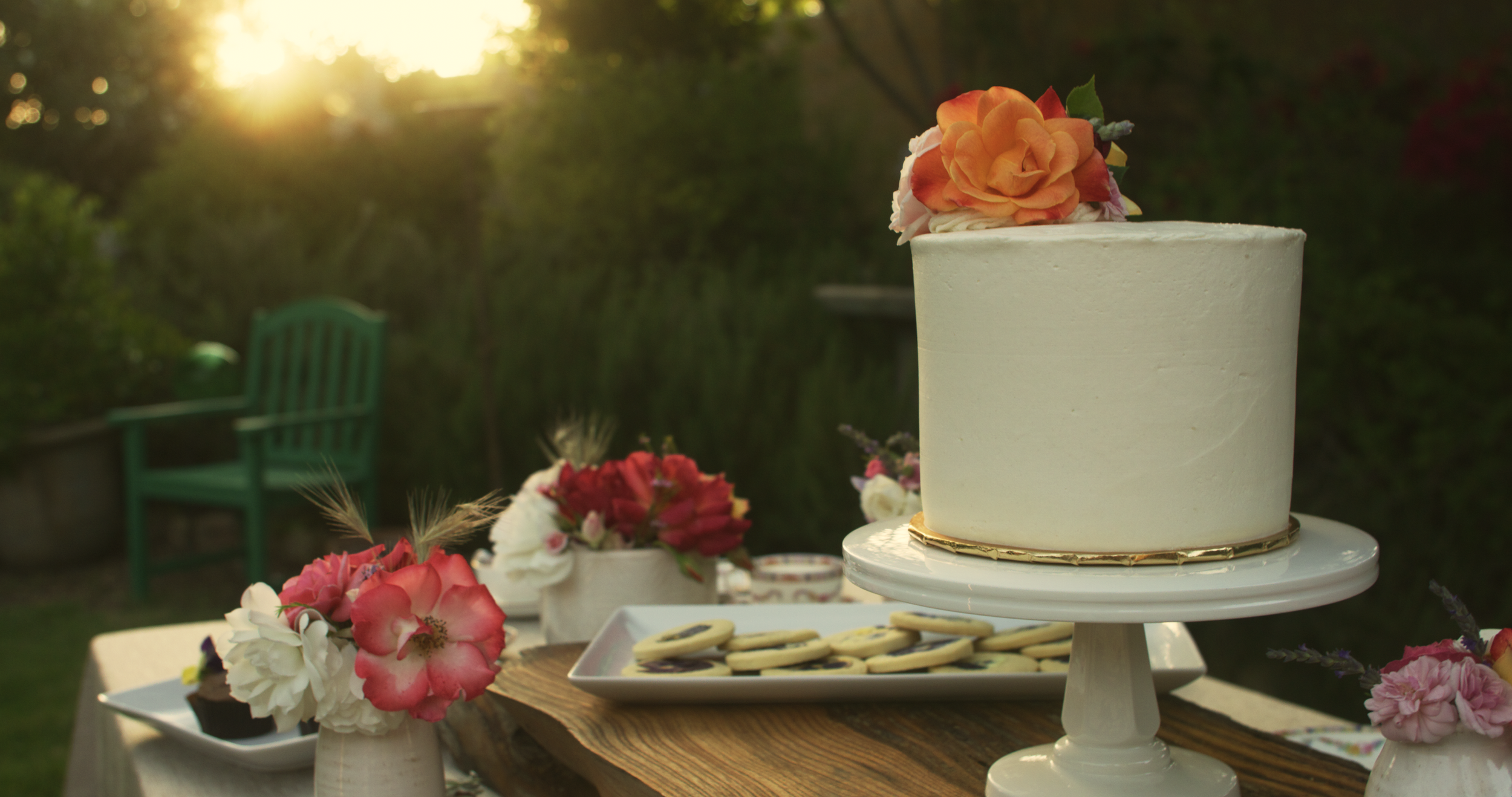 A white cake with a floral decoration on top, set on a white cake stand outdoors with a sunset in the background. The table has flower arrangements and cookies.