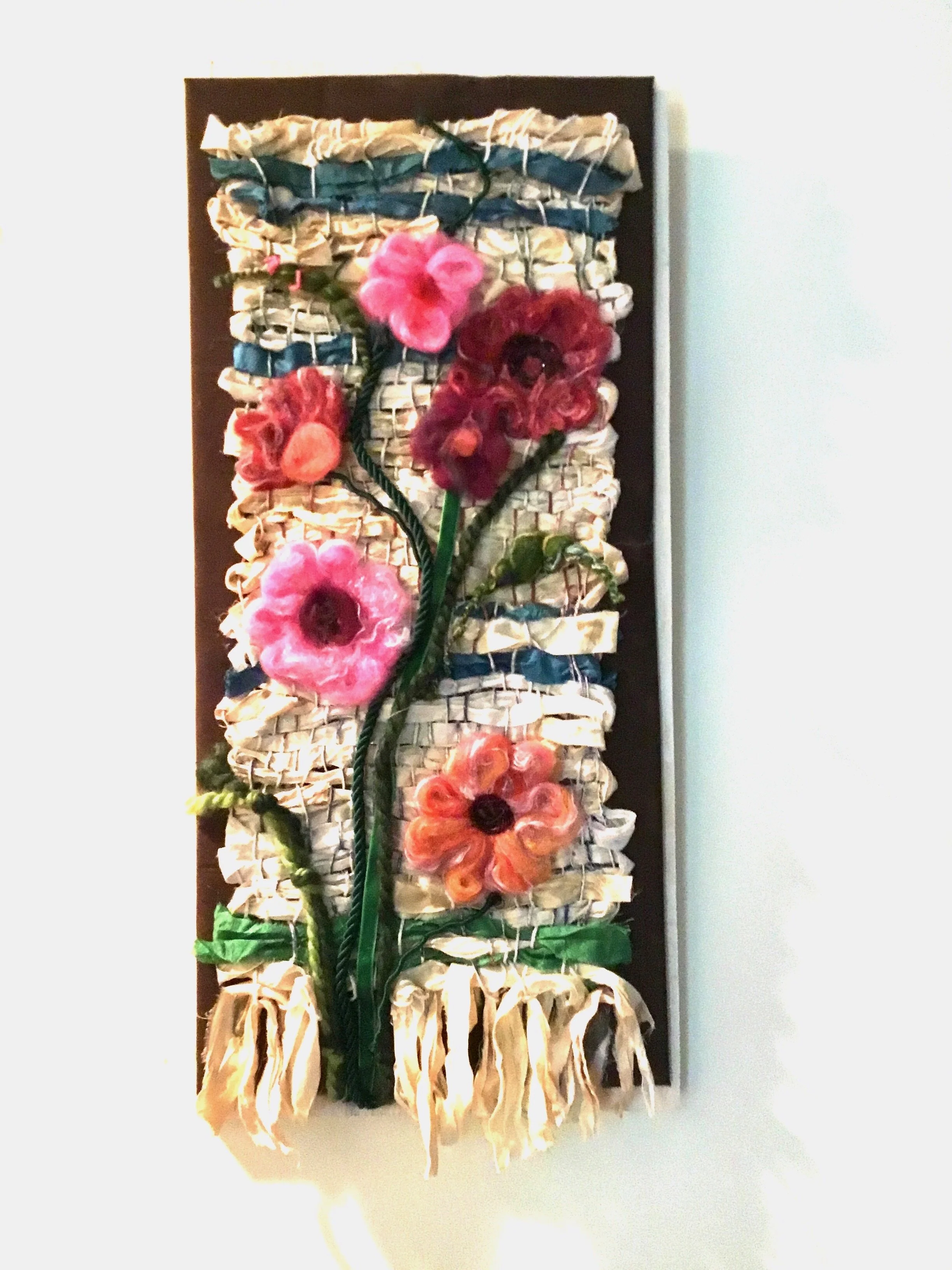 Mixed media wall art featuring fabric, ribbon, and felt flowers arranged to resemble a bouquet of colorful flowers on a woven background.